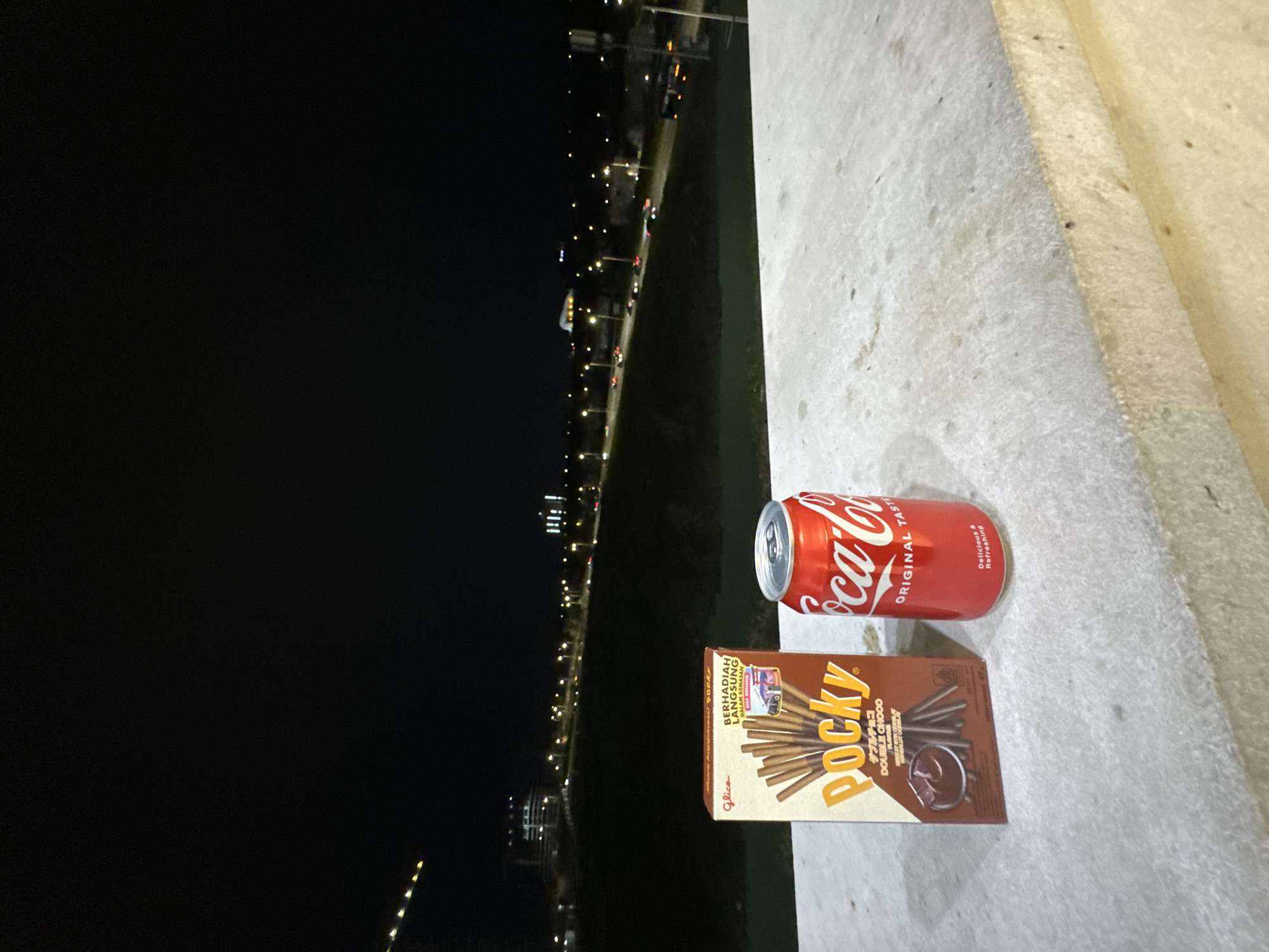 A can of Coca-Cola and a box of Pocky sit on a concrete surface at night. A string of lights, possibly streetlights or traffic, stretches into the distance. The location is not discernible from the image, so a definitive determination of Yerevan, Armenia, is not possible.