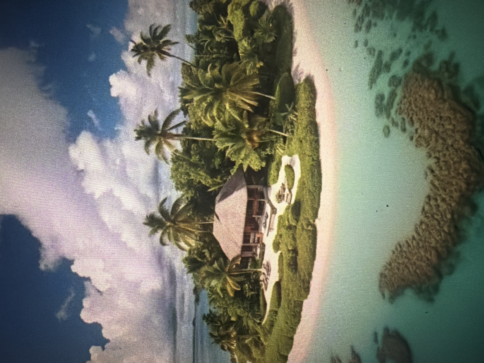 This is an aerial, eye-level, medium shot of a tropical island in the city of Baan Mae Nam, Thailand. The island is lush with green vegetation and dotted with palm trees. A light-colored sandy beach encircles the island, and the clear turquoise water is visible in the foreground, revealing coral formations beneath the surface. A thatched-roof building, likely a villa or resort, is situated on the island, with lounge chairs visible on the patio. The sky above is a vibrant blue with fluffy white clouds. There are no people or animals visible in the image.