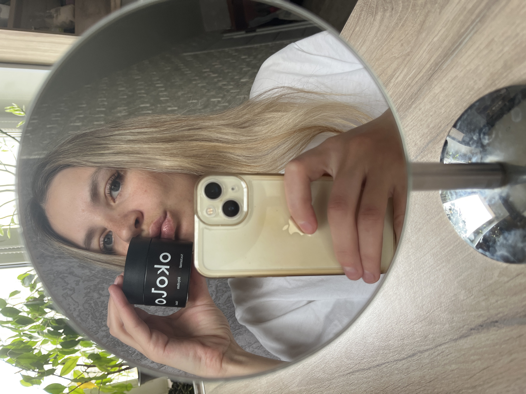 A fair-skinned person with light blonde hair is taking a selfie in a round mirror. The person is wearing a white shirt and holding a black jar labeled "OKOLO" with text in Russian. They are also holding a gold-colored iPhone. The mirror is placed on a light brown wooden surface, possibly a table. The reflection in the mirror shows a blurred background with a window and a plant. The location is Chaikovsky, Russia.