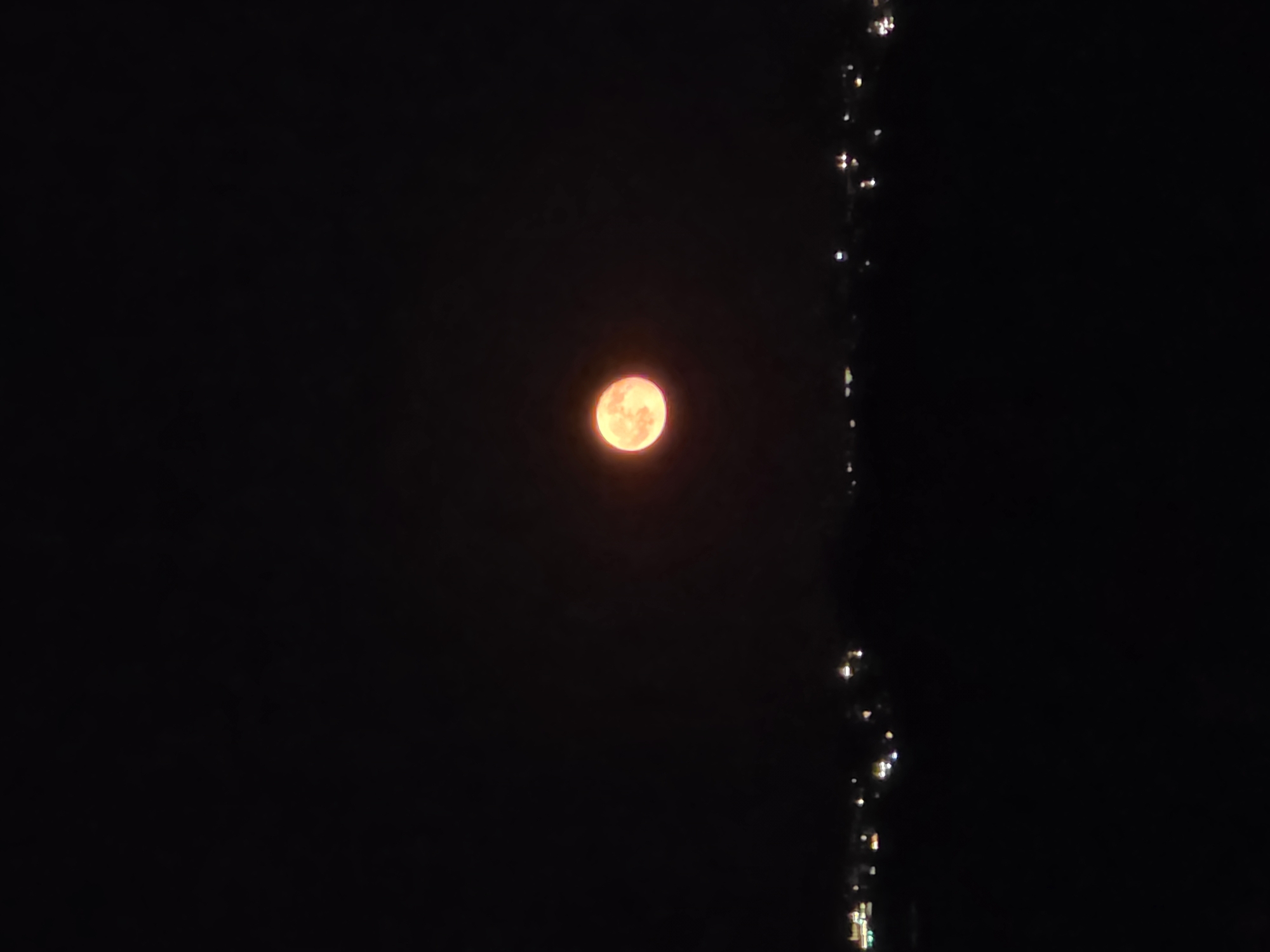 A full, orange-hued moon hangs in the dark night sky above the dimly lit skyline of Chernomorets, Bulgaria. The lower portion of the image depicts a dark, indistinct landscape, likely the ground or distant hills, with a faint line of lights indicating the presence of buildings or streetlights within the city. The moon is the central focal point, casting a soft, warm glow that is slightly diffused by the atmosphere.
