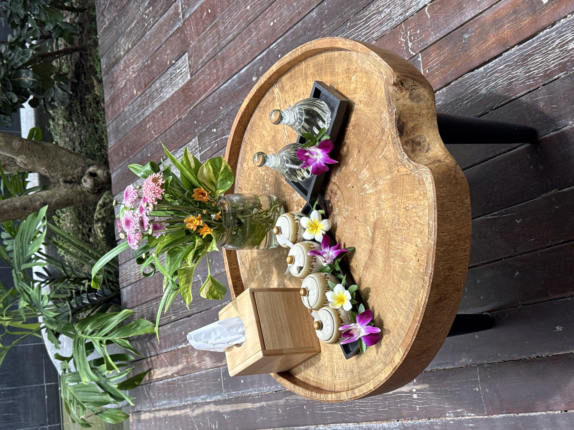 A round, natural wood coffee table with black legs is situated on a weathered wooden deck in Ko Samui, Thailand. On the table, a light wooden tissue box dispenses a single white tissue. To the right of the tissue box is a black tray holding four small, ornate ceramic containers with gold lids, and adorned with purple and white orchids. Further to the right, another black tray holds two clear glass bottles with stoppers. A glass vase filled with a bouquet of pink and orange flowers and greenery sits behind the tissue box. Lush green plants and a tree are visible in the background, suggesting an outdoor setting. The scene appears to be set up for relaxation or a spa experience.