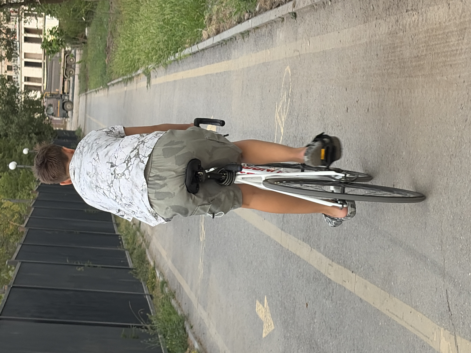 An over-the-shoulder, medium shot shows a young male, seen from the back, riding a white bicycle on a paved bike path in an urban environment. The individual wears a white shirt with a marble-like pattern and camouflage-patterned shorts, along with grey sandals. The bike path is marked with yellow lines and arrows, and is bordered by a grassy area and a tall, dark fence on the left, and a curb with grass on the right. In the background, a brown dump truck is visible on a road, alongside a building with multiple windows. The scene appears to be in a park or designated recreational area within a city.
