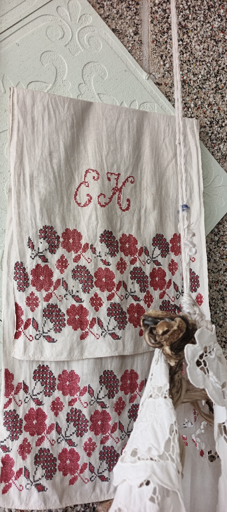 The image shows a piece of embroidered linen hanging against a textured wall. The linen is off-white and features a red and grey floral design along the lower portion, with the embroidered initials "EK" near the top. A piece of white lace and a dried botanical element (possibly a flower or seed head) are hanging in front of the linen on a string. The wall behind the linen is a light green patterned surface and a speckled wall. There is no specific indication of location in the city of Кемуль, Russia.