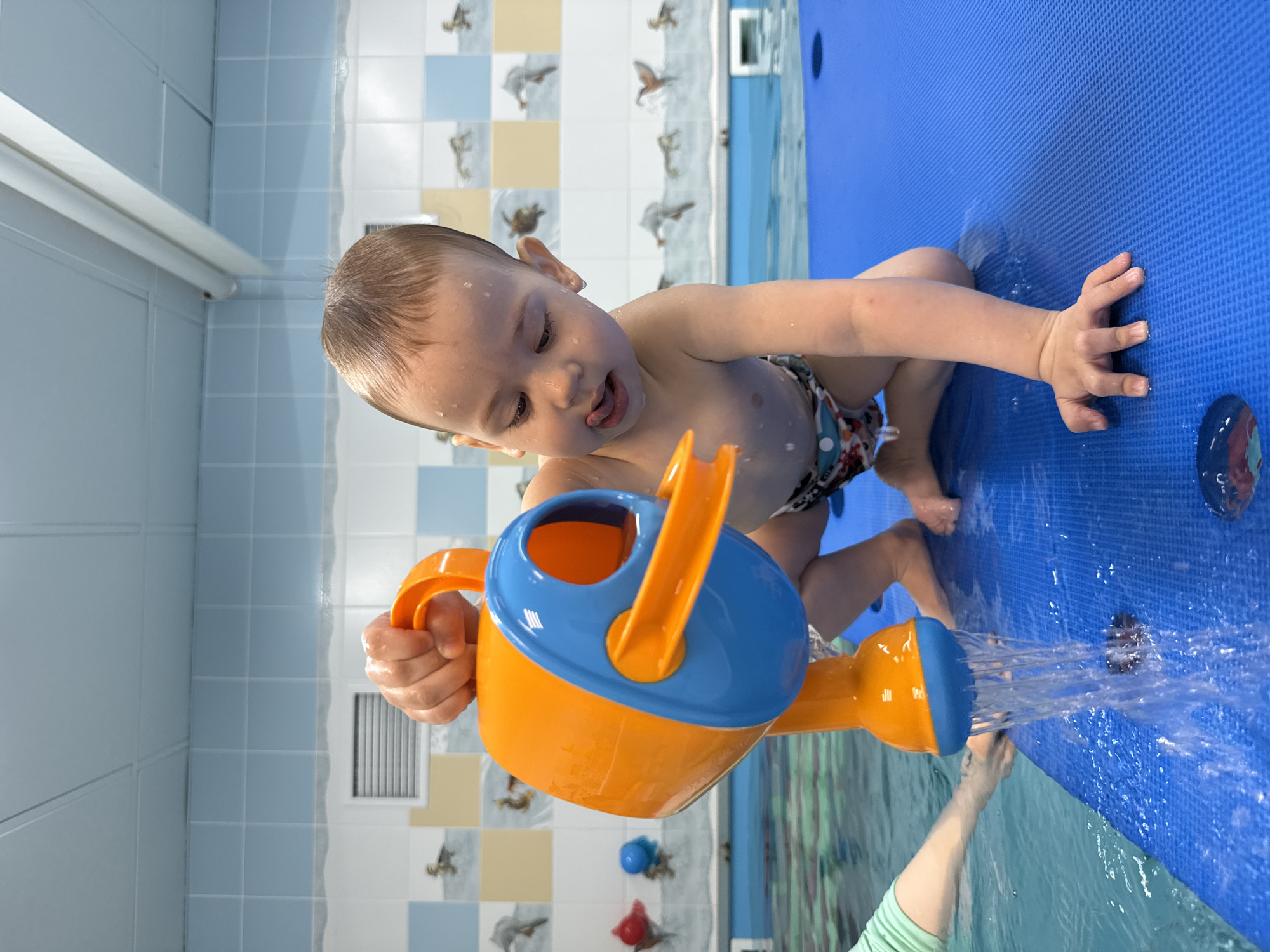 A toddler is kneeling on a blue, cushioned mat in a room with light blue and white tiled walls with images of ducks. The child is holding a blue and orange watering can and pouring water onto the mat. The person assisting the child is visible in the bottom right of the image, presumably a parent. This appears to be an indoor swimming pool. The location is unknown.