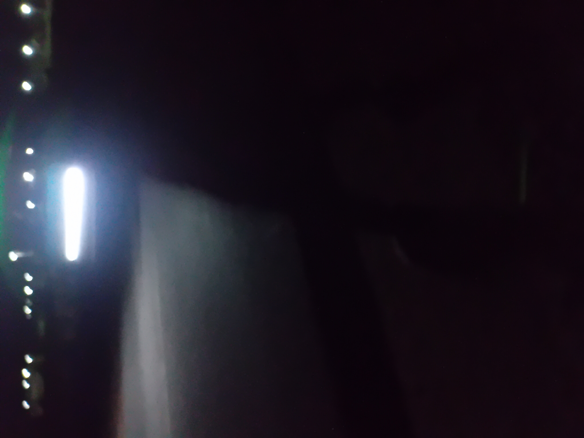 A blurred, dark image depicts a nighttime scene, possibly on a road or open area, with several bright lights visible in the distance. The most prominent light source is a horizontal, glowing white beam situated in the middle left of the frame, suggesting it could be headlights from a vehicle. To the upper left, a series of smaller, circular lights are scattered, appearing as points of light against the dark background, possibly streetlights or other distant illumination. The foreground is almost entirely black, with some hazy, lighter areas suggesting the ground. The overall impression is one of low visibility and artificial light sources in a nocturnal environment.