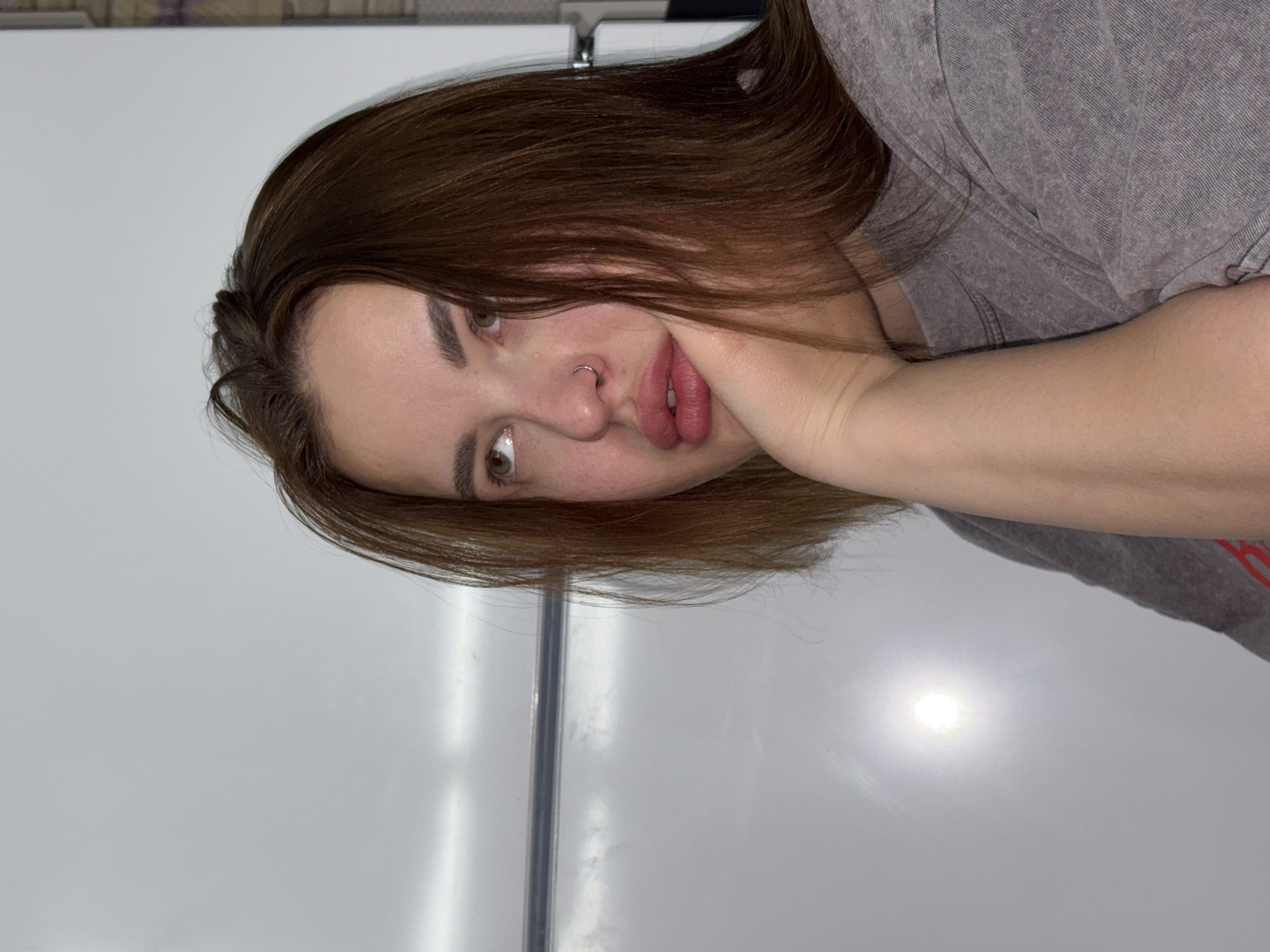 A young woman with light brown hair and fair skin is shown. She has a nose ring and is wearing a gray t-shirt. Her left arm is bent, and her hand is supporting her face. The background is a white surface with a vertical metallic strip. The location is unspecified.