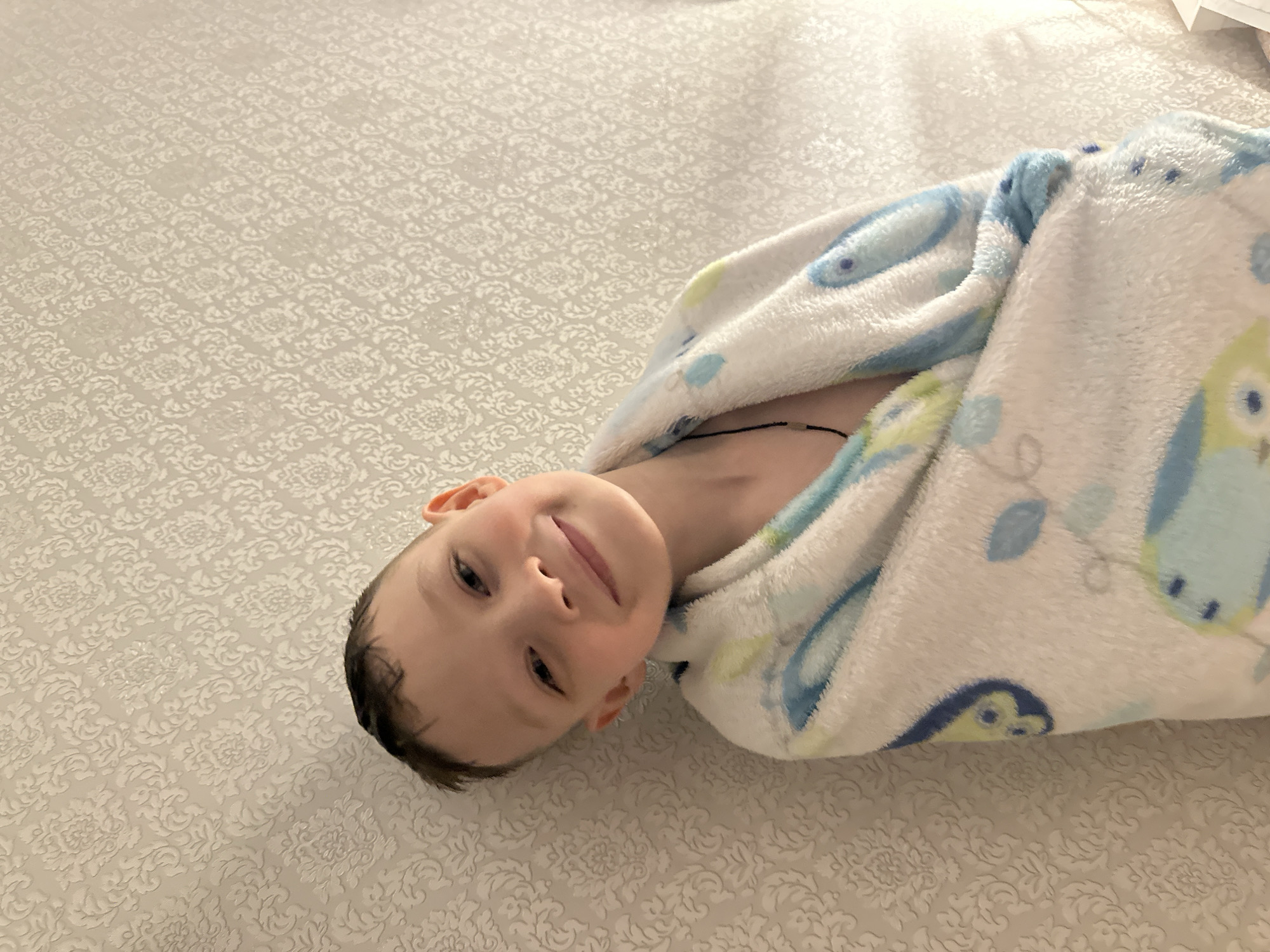 A young boy, wrapped in a white blanket patterned with blue and green owls and leaves, is lying on a light-colored patterned surface. He is facing the camera with a slight smile. His hair is dark and wet. The location is indeterminate, but it is specified as Lyubertsy, Russia.