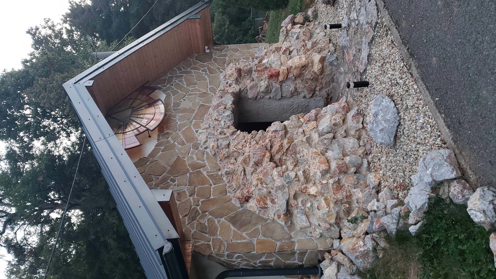 The image depicts the exterior of a building with a distinctive architectural style, seemingly a rustic or fantasy-themed structure. The facade is primarily constructed from irregularly shaped, light brown stones, with a rough-hewn texture. A prominent feature is a semi-circular archway made of larger, reddish-brown and grey rocks, embedded in the stonework, creating an impression of an ancient entrance or cave. This archway leads to a dark opening, presumably a doorway, with a textured surface visible inside.
Above the stone archway, the building's wall transitions to larger, more uniformly shaped stones. On the upper part of the facade, under the eaves of the roof, there is a circular stained-glass window or decorative element. This element has radiating segments in shades of yellow, orange, and red, resembling a sun or a fan. The roof itself is metal, with a dark grey color and visible fasteners. The eaves are lined with wooden planks.
The surrounding environment consists of a gravel path with scattered larger stones, some of which are light grey. To the left of the entrance, there is a patch of green grass with smaller rocks. Two black, cylindrical outdoor lights are visible along the gravel path. In the background, mature trees with dense green foliage dominate the scene, suggesting a natural or semi-rural setting. The sky is overcast or twilight. The image appears to be taken outdoors during the day, possibly in the late afternoon or early evening, given the lighting.
While the image itself does not contain explicit geographical markers, the prompt requests its location in the city of Prešov, Slovakia. Without further information or context, it is not possible to definitively place this specific building within Prešov. However, based on the visual cues of the natural surroundings and the building's unique design, it could be a private residence, a tourist attraction, or a commercial establishment in a less urbanized area of the city or its outskirts.