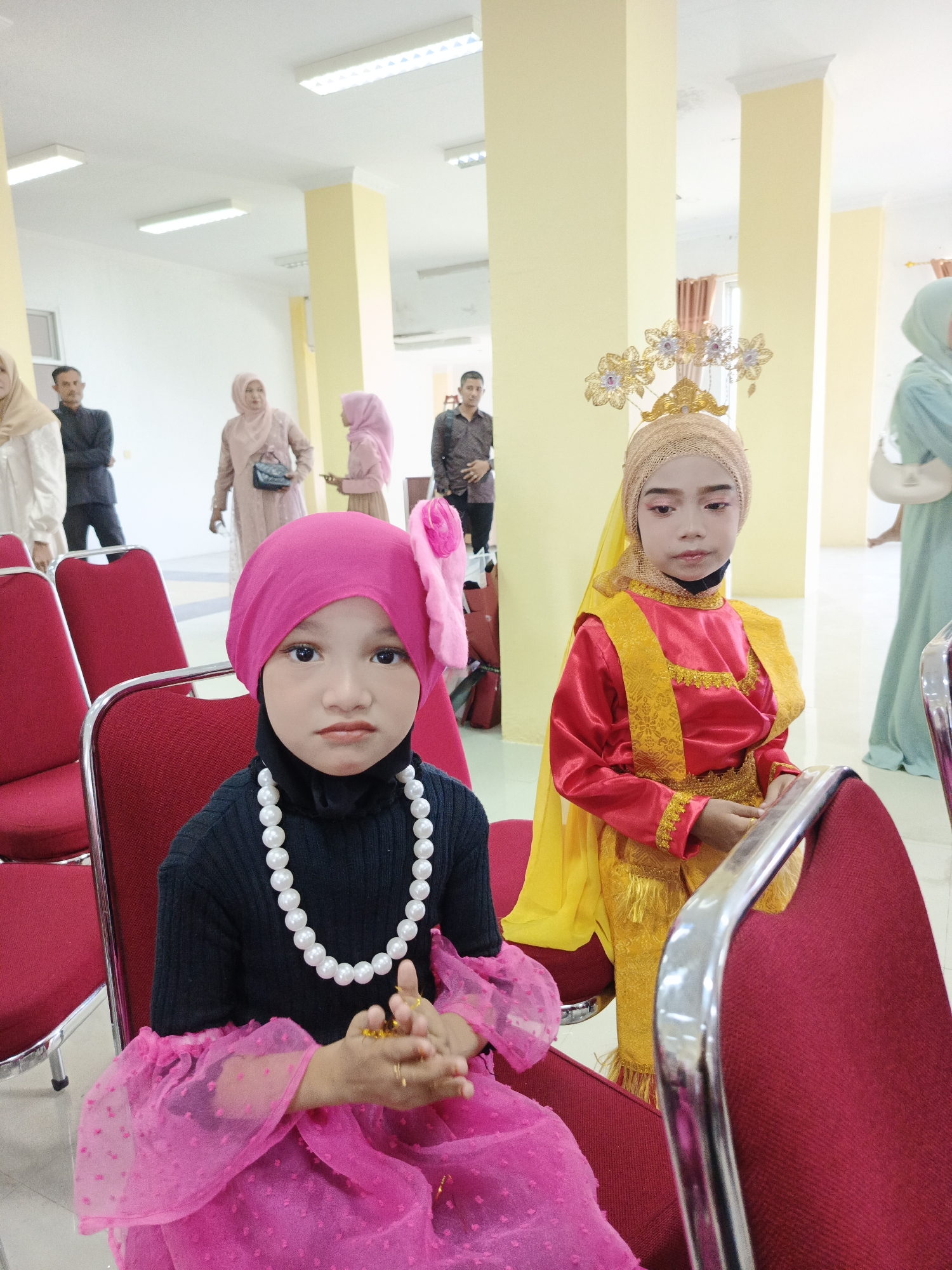 The image depicts an indoor scene, likely a hall or a waiting area, with several people present. In the foreground, two young girls are seated on red chairs, dressed in traditional attire. The girl on the left wears a pink hijab, a black top, and a voluminous pink skirt with sheer sleeves adorned with small polka dots. She is also wearing a pearl necklace. The girl on the right is dressed in a red and gold traditional outfit, complete with a gold headdress. Her makeup is prominent, with bold eyeshadow and lipstick. Several other individuals, appearing to be adults, are visible in the background, some standing and others seated. They are dressed in various casual and formal attire, with some women wearing hijabs. The environment appears to be well-lit with fluorescent ceiling lights and features yellow-painted columns. The overall impression is of a gathering or an event, possibly a cultural celebration or performance, taking place in a public indoor space. While the specific location in Lhokseumawe, Indonesia, cannot be definitively determined from the image alone, the cultural attire and architectural style are consistent with settings in that region.