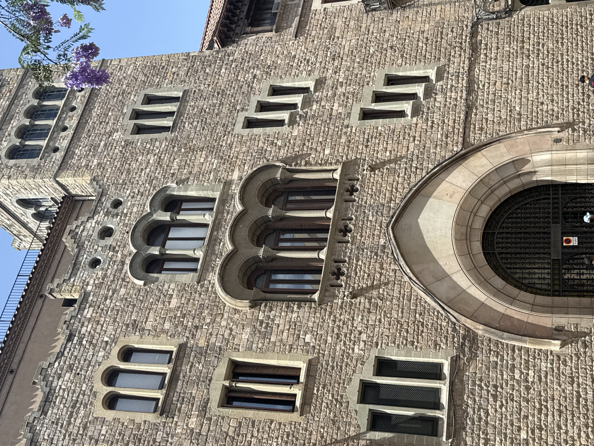 The media file captures a low-angle, upward view of a stone building in Barcelona, Spain, under a clear blue sky. The building's facade is constructed from small, irregularly shaped stones, giving it a textured appearance.
Several windows are visible, exhibiting varied architectural styles. Some are arched with decorative stonework, featuring multiple panes. Others are rectangular with a more modern, minimalist design, some appearing to have blinds or louvers. A large, circular window with intricate metalwork is a prominent feature on the right side of the building.
A branch with vibrant purple flowers, likely wisteria, hangs into the frame from the upper left corner, adding a touch of natural color against the stone. The sunlight casts shadows, indicating it is likely daytime, possibly late morning or early afternoon. Two individuals are faintly visible near the bottom right, suggesting the building might be a public space or has pedestrian access. The overall impression is one of historical architecture bathed in sunlight. No visible text is decipherable in the provided crops.