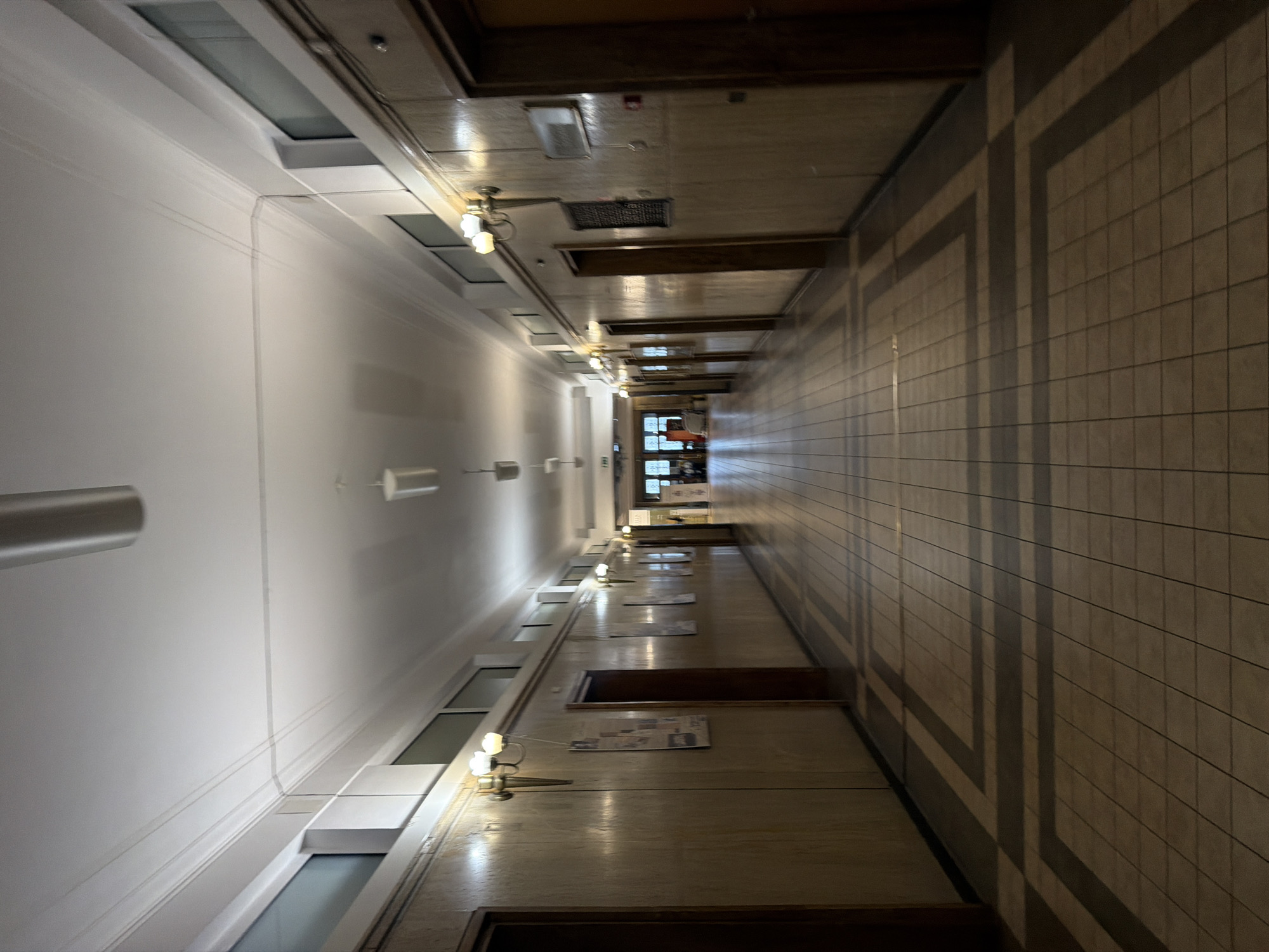 An interior photograph captures a long, empty hallway, likely within a public or institutional building in Sofia, Bulgaria. The perspective is down the length of the corridor, emphasizing its depth and symmetrical elements.
The hallway features a polished, light-colored tiled floor with a recurring pattern of darker, geometric outlines. The high walls flank this central path. The left wall is painted white, adorned with decorative molding near the ceiling, and integrates a series of recessed rectangular windows or transom openings that allow some ambient light. The right wall is covered in light, rectangular tiles and is punctuated by multiple wall-mounted brass sconces, each holding several bulbs, providing artificial illumination. Between these light fixtures, various framed documents or pictures are displayed, though their content is not legible.
The ceiling is composed of light-toned wooden panels interspersed with darker wooden beams and incorporates recessed lighting elements and air vents. Strong, geometric shadows are cast diagonally across the tiled right wall and partially on the floor, suggesting external light sources from unseen windows or architectural features outside the immediate frame. At the far end of the hallway, a brighter area is visible, indicating another set of doors or an opening to a different section of the building. The absence of people and the architectural style evoke a quiet, possibly historical, and formal atmosphere. No specific text is discernible.