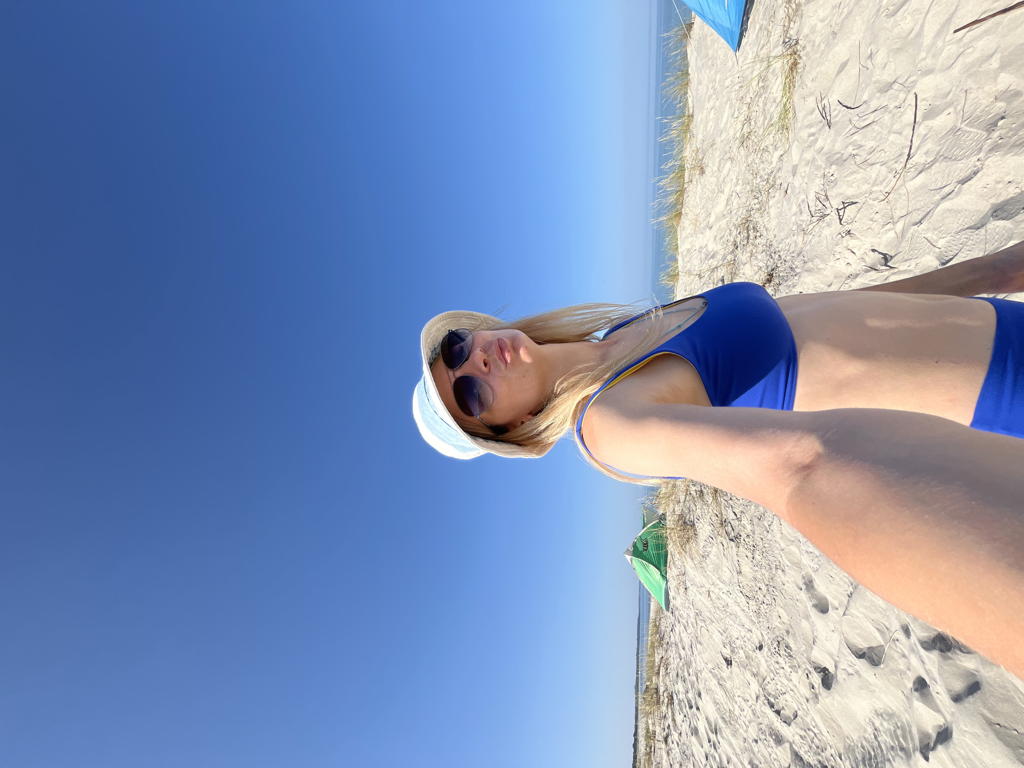 A medium shot captures a blonde woman with long hair, wearing a blue bikini, a white and blue bucket hat, and sunglasses, as she takes a selfie on a sandy beach. She is puckering her lips and looking towards the camera. Behind her, two tents are visible, one green and white on the left and a partially visible blue one on the right. The background consists of sand dunes with sparse dry grass, and the blue ocean meeting the clear blue sky. The setting appears to be a beach in Okunevo, Russia.