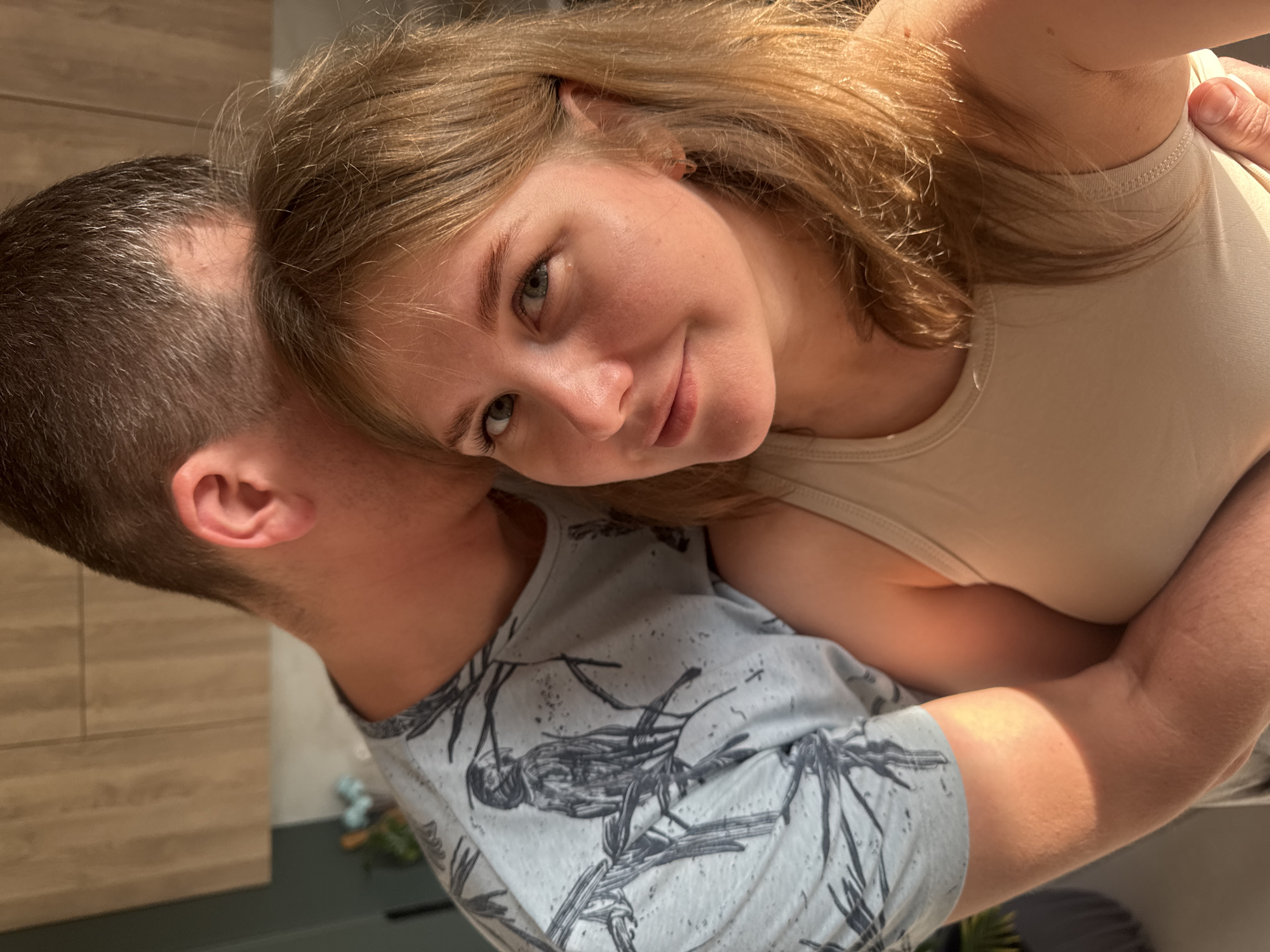A woman with light brown hair is embracing a man with short, dark hair. She is wearing a light beige tank top, while the man is wearing a gray t-shirt with a bird design. They appear to be indoors in a domestic setting, perhaps a kitchen, with light wood cabinets and dark green countertops visible in the background. The location is Ферма, Russia.