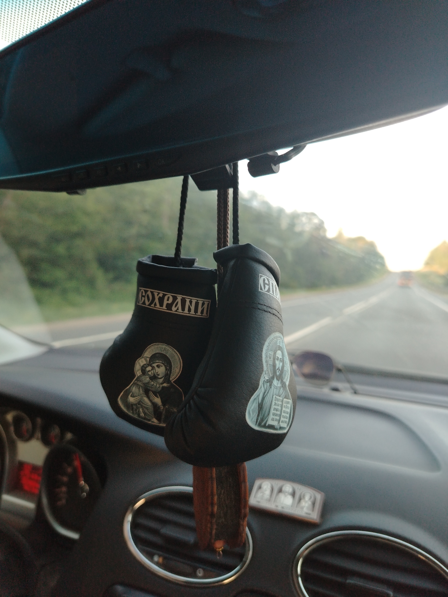 The image shows the interior of a car, specifically the dashboard and rearview mirror area. Hanging from the rearview mirror are a pair of small, black boxing gloves. One glove has an image of the Virgin Mary and the child Jesus, while the other has an image of Jesus Christ. The word "Сохрани" (Save) is written on one glove. The dashboard features air vents with silver trim and a small religious icon. Outside the car, a road stretches into the distance, lined with trees. The location is Новое, Russia.
