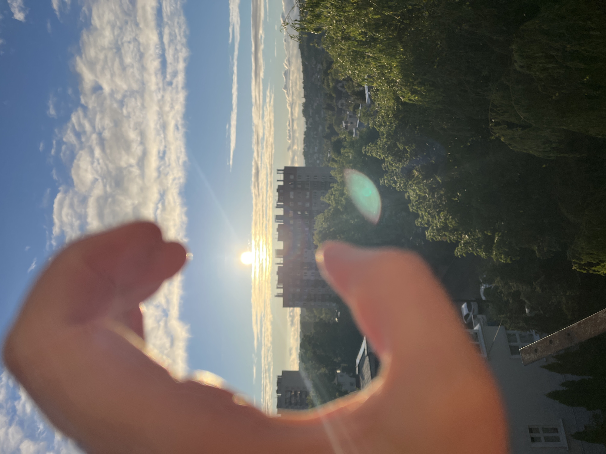 The image is a well-lit outdoor shot capturing a human hand in the foreground, forming an incomplete heart or 'C' shape, with the sun prominently framed within its curve. The hand, significantly out of focus, is bathed in warm, golden light from behind, highlighting its edges with a soft glow and showing a warm skin tone.
In the midground, the sun is a brilliant, circular orb, radiating intense light and producing visible lens flares, including a distinct greenish-blue circular artifact on the lower right and bright linear streaks. Directly beneath the sun, a multi-story building with a red-brown roofline and numerous windows stands centrally, framed by the hand. To the left of the sun, partially obscured by the hand, another lighter-colored, multi-story building is visible.
Below the buildings and extending into the lower half of the frame is a dense canopy of lush green trees, illuminated by the low sun, creating areas of bright highlights and deep shadow. A thin, dark, out-of-focus linear object, possibly a railing or structural beam, diagonally crosses the bottom-center of the image, behind the lower part of the hand.
The background reveals a clear blue sky with scattered white cumulus clouds, forming distinct horizontal bands. Beyond the immediate trees and buildings, a sprawling landscape of distant hills or higher ground is visible, dotted with more trees and hints of urban development. The overall lighting suggests either sunrise or sunset, indicative of the "golden hour."
The location is in the city of Unknown, Unknown.