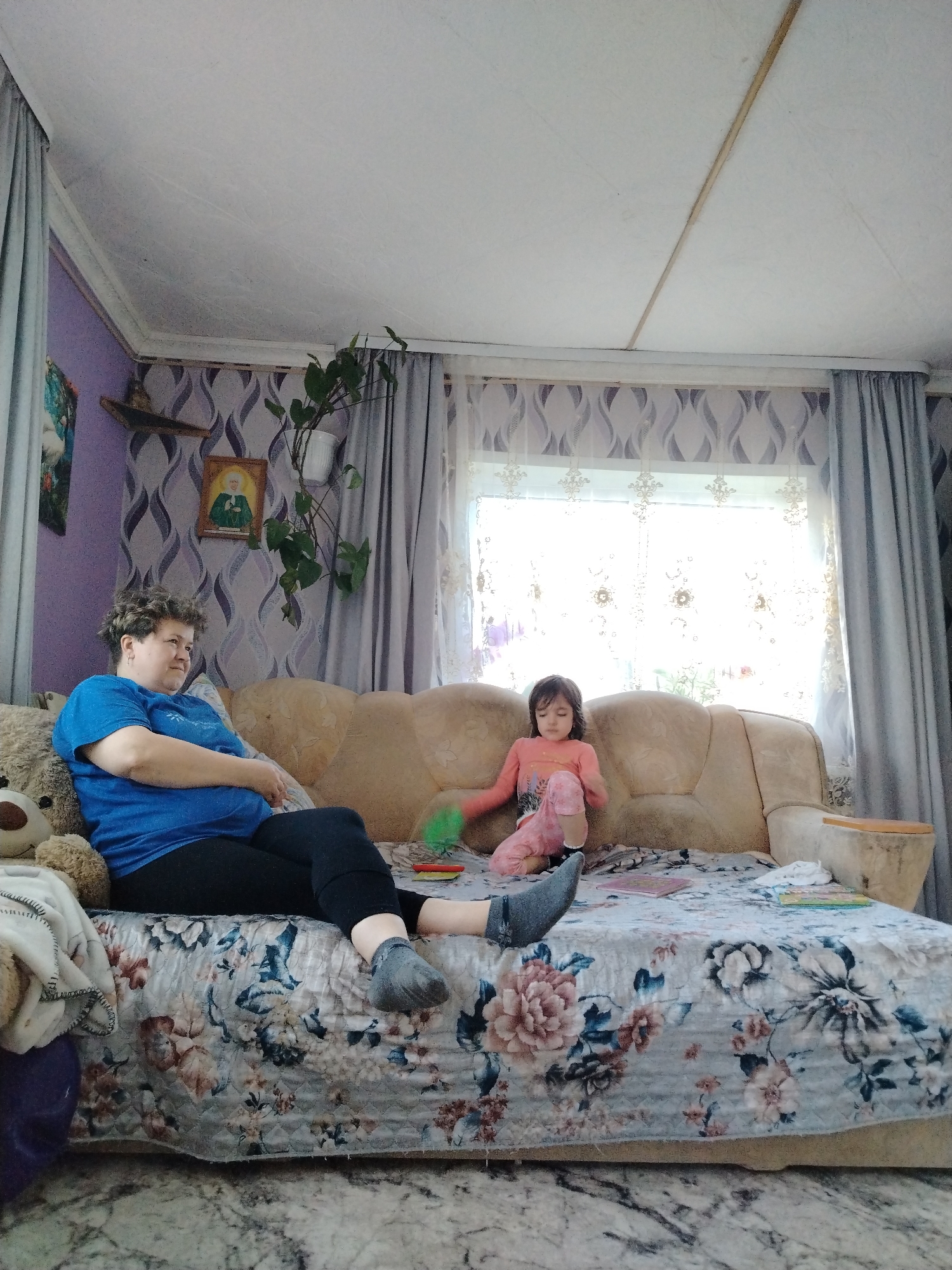 The image shows a woman and a young child sitting on a couch in what appears to be a domestic setting in the city of Гожан, Russia.
The woman is seated on the left side of the couch, wearing a blue t-shirt and black leggings. She has short curly hair and her arms are crossed. Next to her on the couch is a large teddy bear.
A young child is sitting on the right side of the couch, wearing pink clothing. The couch is covered with a floral patterned fabric. There are art supplies on the couch.
Behind the couch is a window with sheer curtains and heavier gray drapes. The walls are decorated with purple wallpaper. A plant is hanging in front of the window. A picture and a small shelf are mounted on the wall to the left of the window. The ceiling is white. The floor has a mottled pattern.