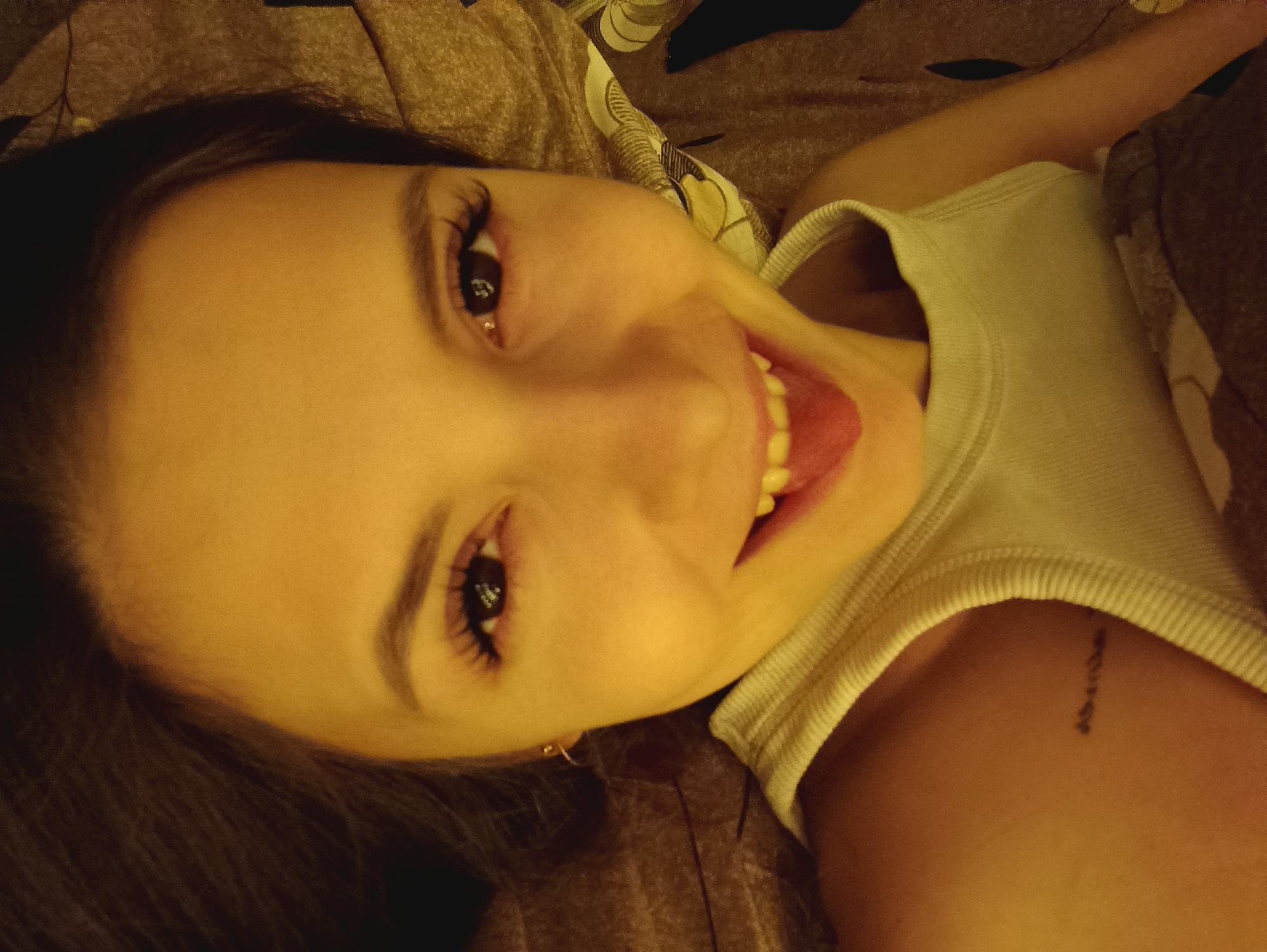 A person is lying on a bed. The person is wearing a white tank top and has dark hair. The person is smiling and sticking their tongue out. There is a tattoo on the person's shoulder. The bed has a brown patterned sheet.