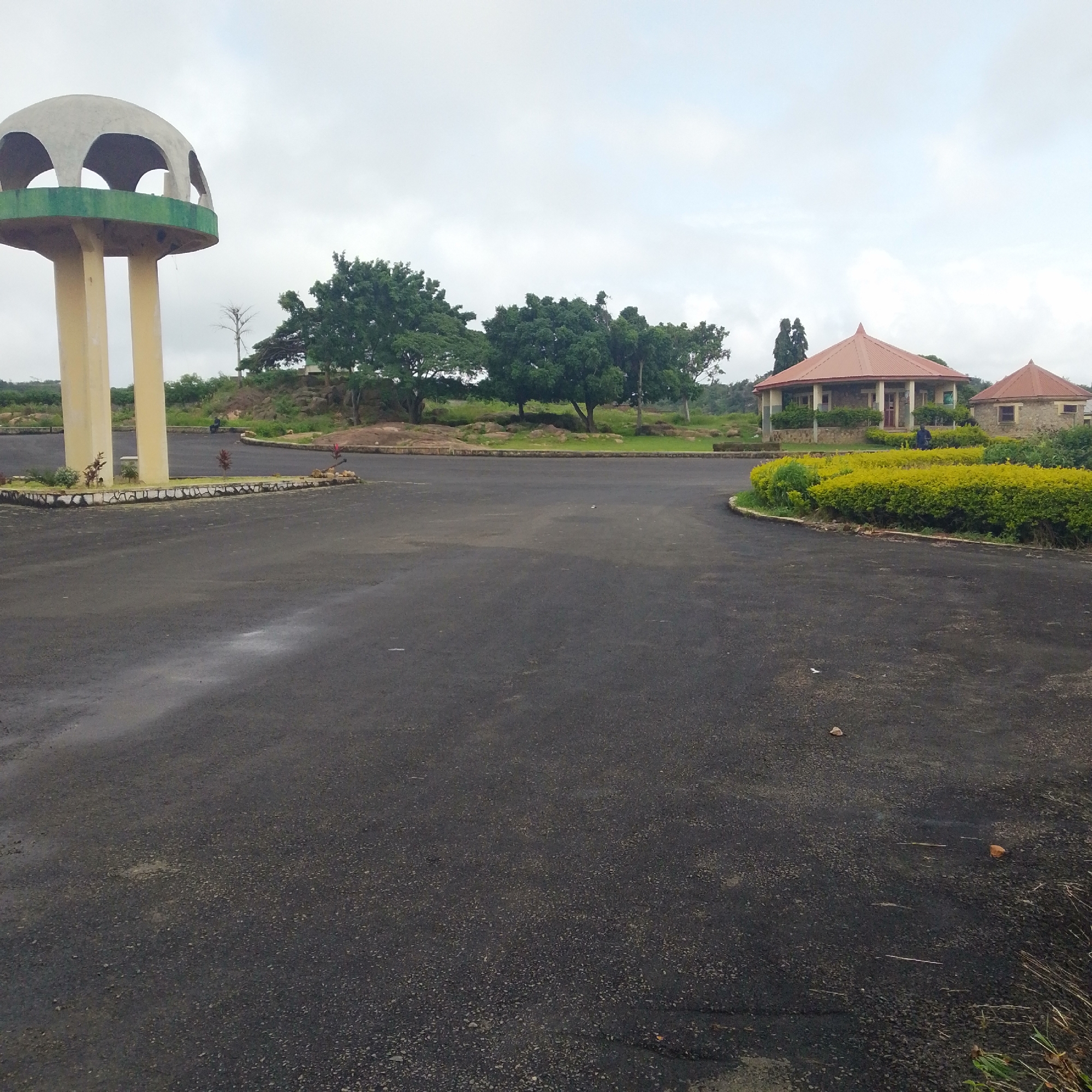 An exterior, wide-angle, eye-level shot captures a paved area in front of two small, stone buildings with conical red roofs, a large water tower, and lush green trees under a cloudy sky. The paved area is dark asphalt with some scattered pebbles and a few small leaves. To the left, a water tower with a dome-shaped top and three arched openings stands on three cream-colored cylindrical supports. A green band runs horizontally around the base of the dome. In the mid-ground, on the right, are the two buildings. The larger one, closer to the foreground, has a red conical roof, white columns supporting a verandah, and stone walls. The smaller building to its right also has a red conical roof and stone walls. A man in a blue shirt and dark pants stands between the two buildings, facing away from the camera. In front of the buildings, a curved, manicured hedge of bright green bushes lines the paved area. Behind the buildings and the water tower, rolling hills are covered with greenery and large rocks. Trees are scattered throughout the landscape. The overall scene is one of a well-maintained outdoor area with architectural elements and natural vegetation.