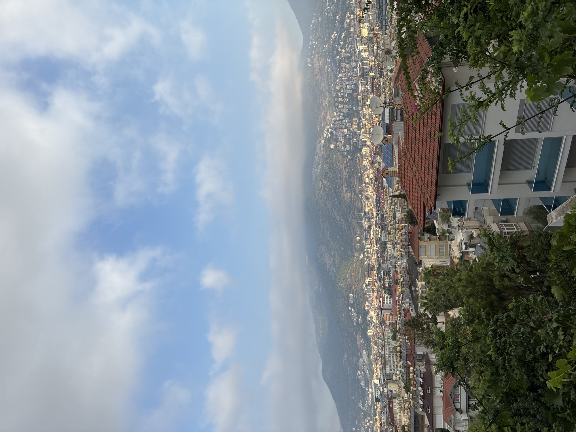 The image depicts a panoramic view of the city of Alanya, Turkey, from an elevated vantage point. In the foreground, lush green foliage of trees and vines partially obscures a modern multi-story building with white walls and red-tiled roofs, featuring blue-tinted glass balconies. Several satellite dishes are visible on a rooftop.
The mid-ground is densely populated with buildings of varying heights and architectural styles, predominantly featuring white and light-colored facades, many with red-tiled roofs and solar water heaters. The city stretches horizontally across the frame, extending towards the foothills of a large mountain range in the background.
The mountain range itself is covered in vegetation and dotted with numerous houses and structures, appearing to ascend towards the peak. The upper portion of the mountains is partially obscured by a layer of soft, white clouds, creating a hazy effect. The sky above is a vibrant blue, with scattered white clouds, some of which are thin and wispy, while others are denser. The overall impression is of a sunny day with some atmospheric haze. The scene is devoid of any visible people or animals.