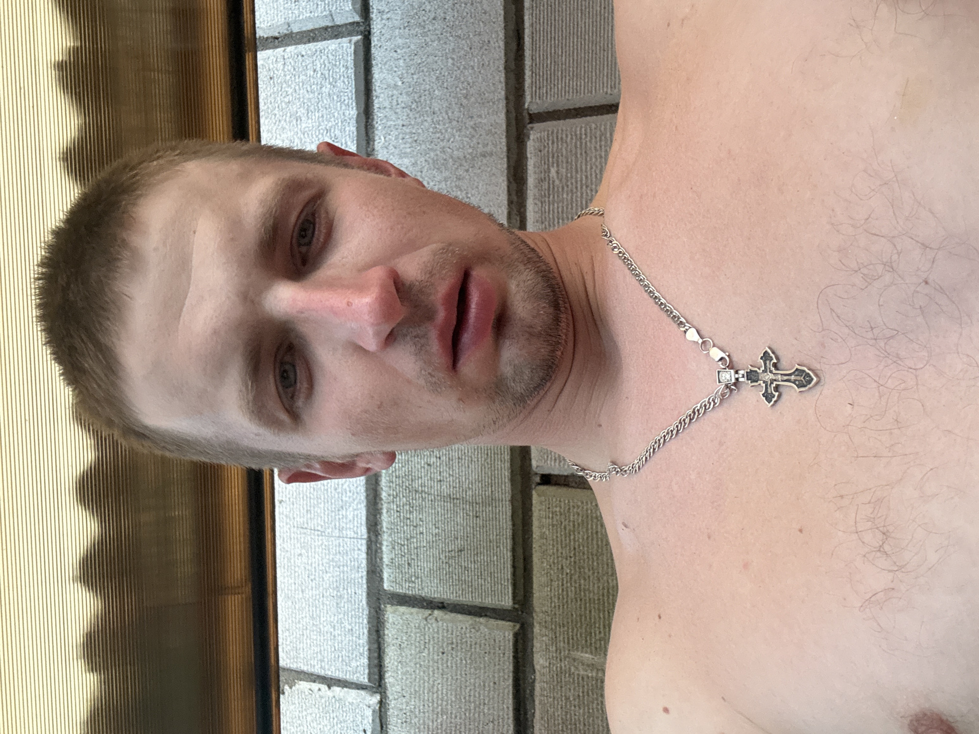 The image is a close-up, waist-up, selfie-style photograph of a fair-skinned Caucasian male, appearing to be in his late 20s to early 30s. He has short, light brown hair, a fair complexion, and light blue or gray eyes. His facial expression is neutral, with his mouth slightly parted. He has some stubble on his chin and upper lip. He is wearing a silver chain necklace with a cross pendant. His chest and shoulders are bare. The background consists of a textured, light-colored brick wall. Above the brick wall, there is a yellowish, ribbed, translucent structure, possibly a canopy or roof. The lighting suggests an outdoor setting. The image was taken in Maladzyechna, Belarus.