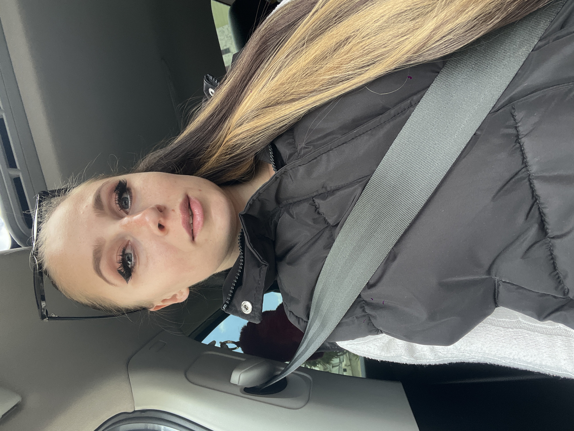 A woman is in a car, wearing a black puffer jacket and a seatbelt. She has long, highlighted hair and distinct eye makeup. Sunglasses rest on her head. The interior of the car is visible, and the scene is bathed in natural light. The image does not show a specific location, so it's impossible to confirm if it is in Budva, Montenegro.