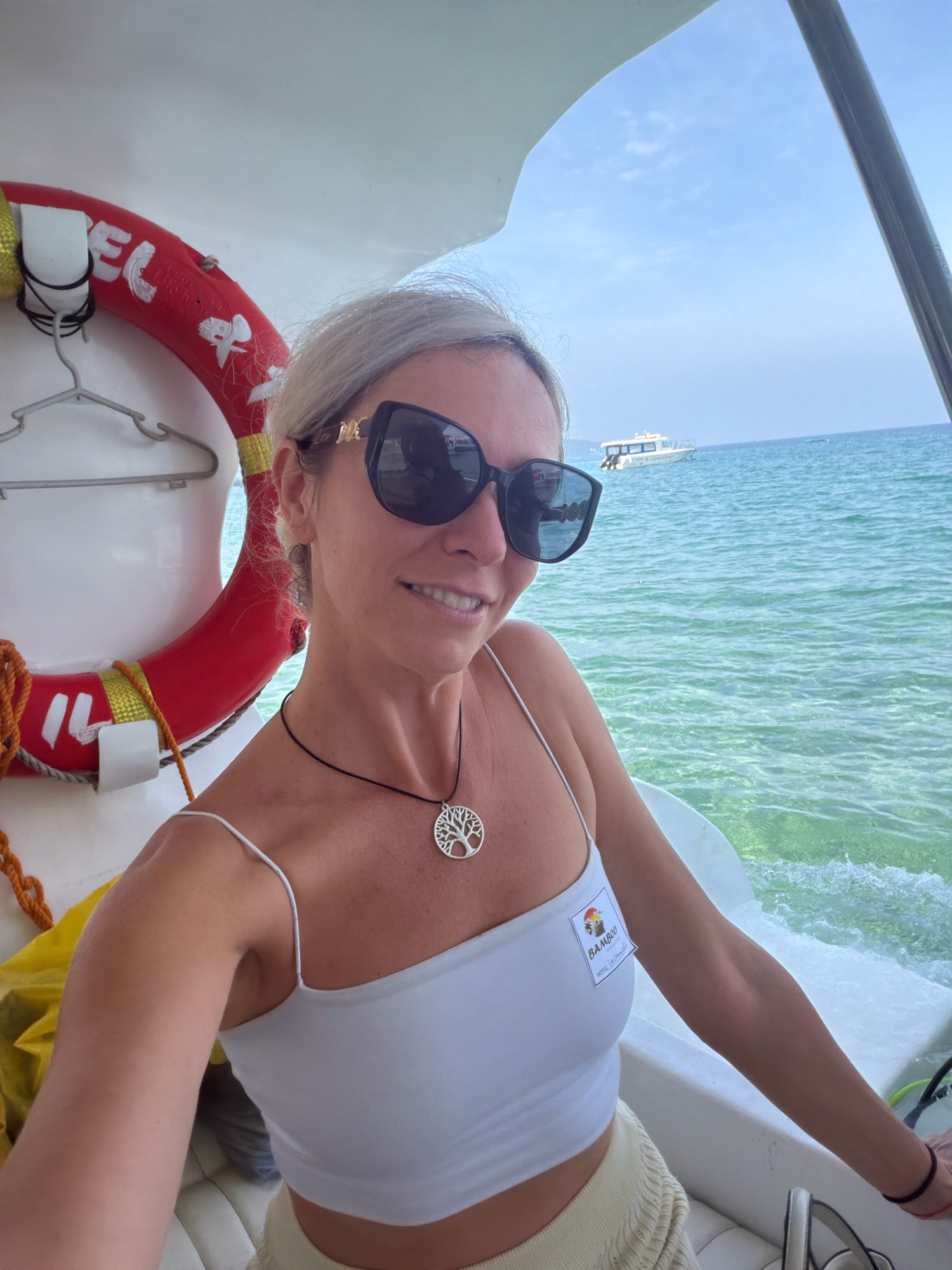 A woman with blonde hair, wearing sunglasses, a white tank top, and a necklace with a tree of life pendant, is taking a selfie on a boat. She is smiling and appears to be holding the steering wheel. In the background, the turquoise ocean is visible, with another boat in the distance. A red lifebuoy hangs on the left side of the boat. A badge with "BAMBOO HOTEL Boracay" printed on it is attached to her top. The setting is consistent with the waters surrounding Boracay Island, in Malay, Philippines.