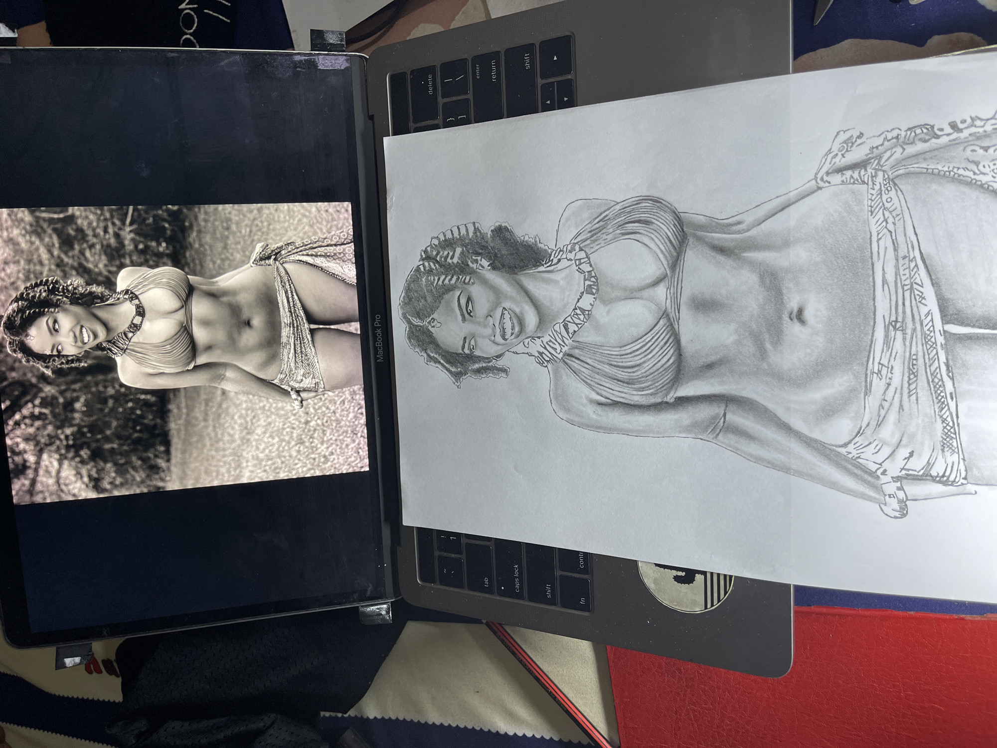 The image shows a laptop displaying a grayscale photo of a woman in a bikini and jewelry. Lying atop the laptop is a pencil drawing that attempts to replicate the displayed photo. The background of the photo on the laptop is blurry, suggestive of an outdoor environment, possibly a garden. The pencil drawing is placed on the keyboard of the laptop, and its orientation matches that of the photograph on the screen. The surrounding environment includes a glimpse of a red surface and some clothing, alluding to a casual, indoor setting. There is no indication of location in the city of Jos, Nigeria.