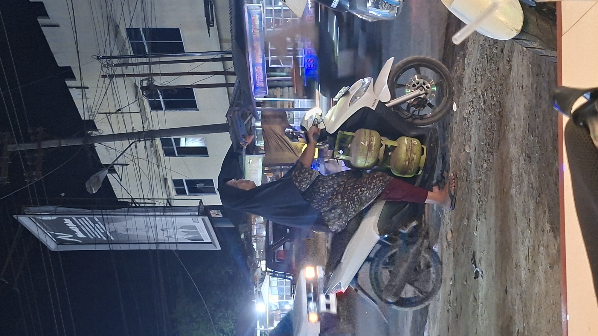 In Keudegeudong, Indonesia, at night, a woman wearing a black hijab and a patterned dress is riding a white Yamaha scooter. She is carrying two green LPG gas cylinders strapped to the back of the scooter. The scooter is on a dirt road in front of a building with several windows and an illuminated sign that reads "ESTIMASI FORM..." The scene is illuminated by streetlights and lights from the building. Other vehicles are visible in the background, including a white car. The woman is looking to her right.