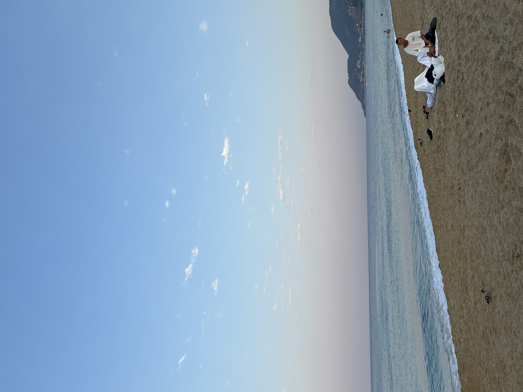 An elevated, eye-level, wide shot captures a serene beach scene in Alanya, Turkey, with the city's iconic coastline visible in the distance. The sky, a gradient of soft blue transitioning to a pale pink near the horizon, is dotted with wispy clouds. The calm sea displays shades of turquoise and blue, with gentle waves lapping at the sandy shore.
In the foreground, a man sits on a blanket on the sand, dressed in a white t-shirt and dark trousers. He appears to be attending to another person who is lying down, covered by a white sheet, with their head resting on the man's lap. The scene suggests a moment of rest or care. Several items are scattered around them, including clothing, shoes, and a plastic bag, indicating a relaxed beach outing. Two pigeons peck at the sand nearby, adding a touch of natural life to the human-occupied space.
Further down the beach, in the middle ground, a few other individuals are visible in the water, swimming or wading. The coastline is framed by a mountainous landscape that slopes down to the sea, where buildings and structures of Alanya are nestled. The overall atmosphere is peaceful and tranquil, characteristic of a late afternoon or early evening at the beach.