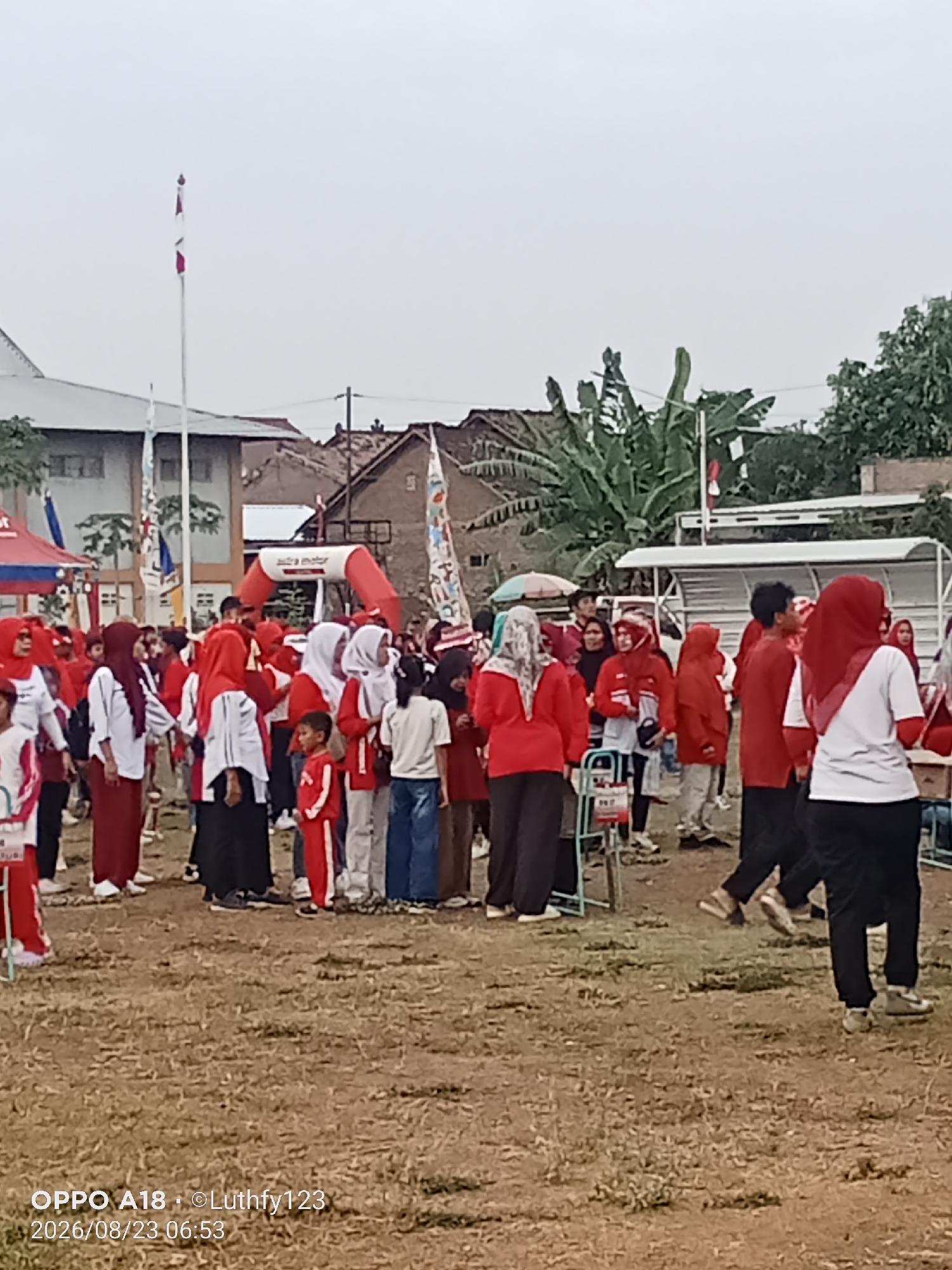 A group of people, primarily children and adults wearing red and white, are gathered outdoors on a grassy field in Cawas, Indonesia. Many are wearing headscarves, and some appear to be participating in an event, possibly a celebration or competition. There are tents and a red archway in the background, suggesting a public gathering. The sky is overcast, and trees are visible in the distance. The image is dated August 23, 2026.