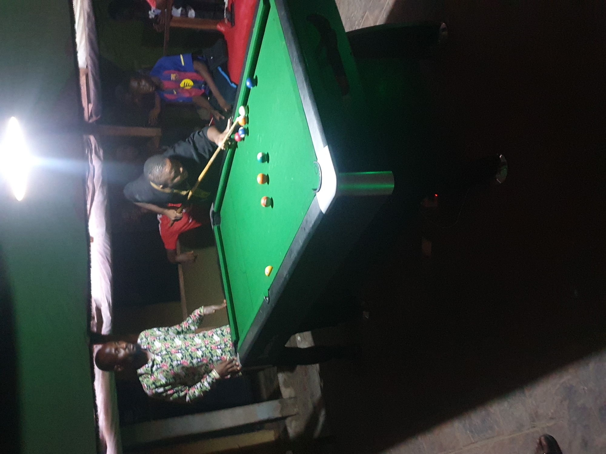 The image captures a nighttime scene inside what appears to be a dimly lit recreational space, possibly a bar or community center, in Lokoja, Nigeria. The central focus is a green-felt pool table where several men are engaged in a game of billiards.
One man, wearing a black t-shirt, is actively playing, leaning over the table with a cue stick in hand, aiming at the balls. His posture suggests concentration on the shot. Several pool balls, including solids and stripes, are scattered across the table's surface.
Behind the player, another man in a blue and red striped t-shirt, resembling a football jersey with a club logo (likely FC Barcelona, indicated by the colors and shield-like emblem, with partially visible letters like 'M L' or 'W L' and an emblem on the chest), stands observing the game. To the lower left of the table, a third man wearing a distinctive light-colored, long-sleeve shirt with a green and white floral or abstract pattern is also watching.
A strong, bright artificial light source, possibly a bare bulb, illuminates the scene from the left, creating a significant glare and high contrast. The surrounding environment is dark, with vague outlines of structures and other individuals visible in the shadows, suggesting other patrons or furniture. The floor appears to be a concrete or tiled surface. The atmosphere seems casual and social, centered around the game. No specific weather conditions are discernible, as the activity takes place indoors or under a cover. Emotions are generally focused and engaged in the game.