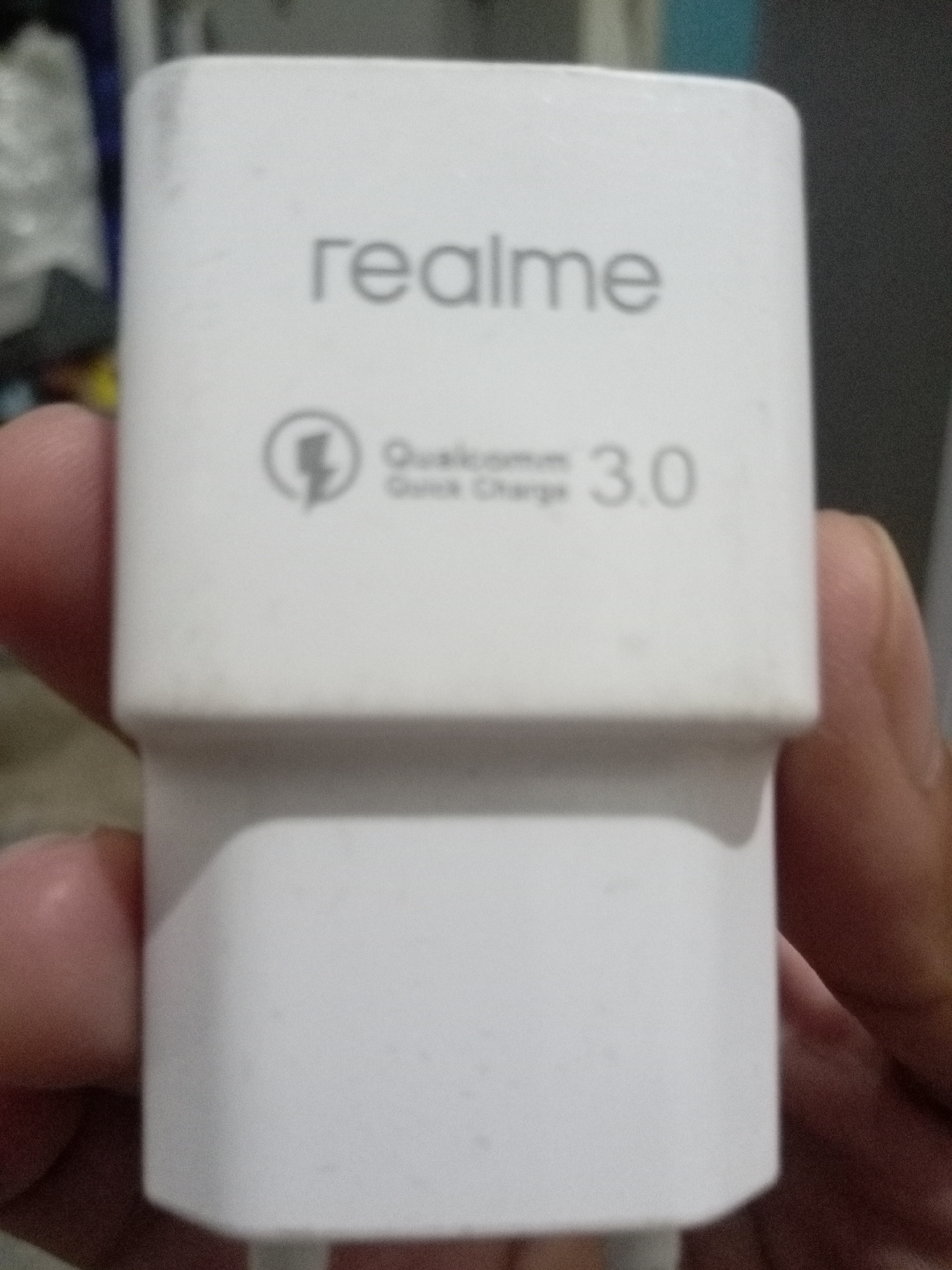 A white Realme charger is held by a person in Batam, Indonesia. The charger has "realme" printed on its front, along with a lightning bolt logo and "Qualcomm Quick Charge 3.0". The image is a close-up shot, with the charger occupying most of the frame. The background is out of focus and suggests an indoor environment.