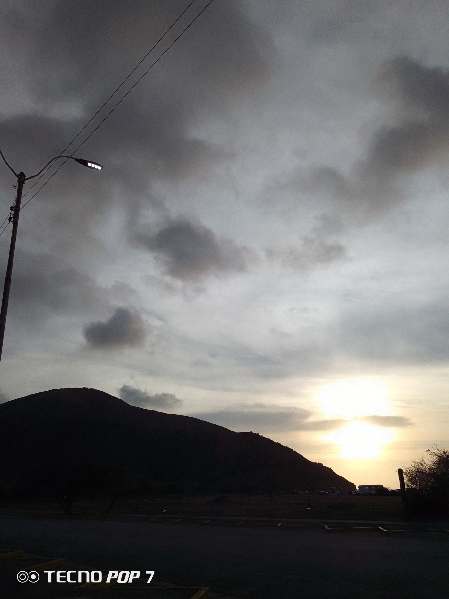 The image captures a dusky scene in Altagracia, Venezuela. The sky is filled with heavy, grey clouds, partially obscuring the setting sun. A silhouette of a dark, mountainous form dominates the midground. In the foreground, there is a paved surface with yellow road markings. A light post with a modern LED fixture stands on the left. A vehicle and foliage are partially visible on the right side of the mountain. The text "TECNO POP 7" appears at the bottom of the image.