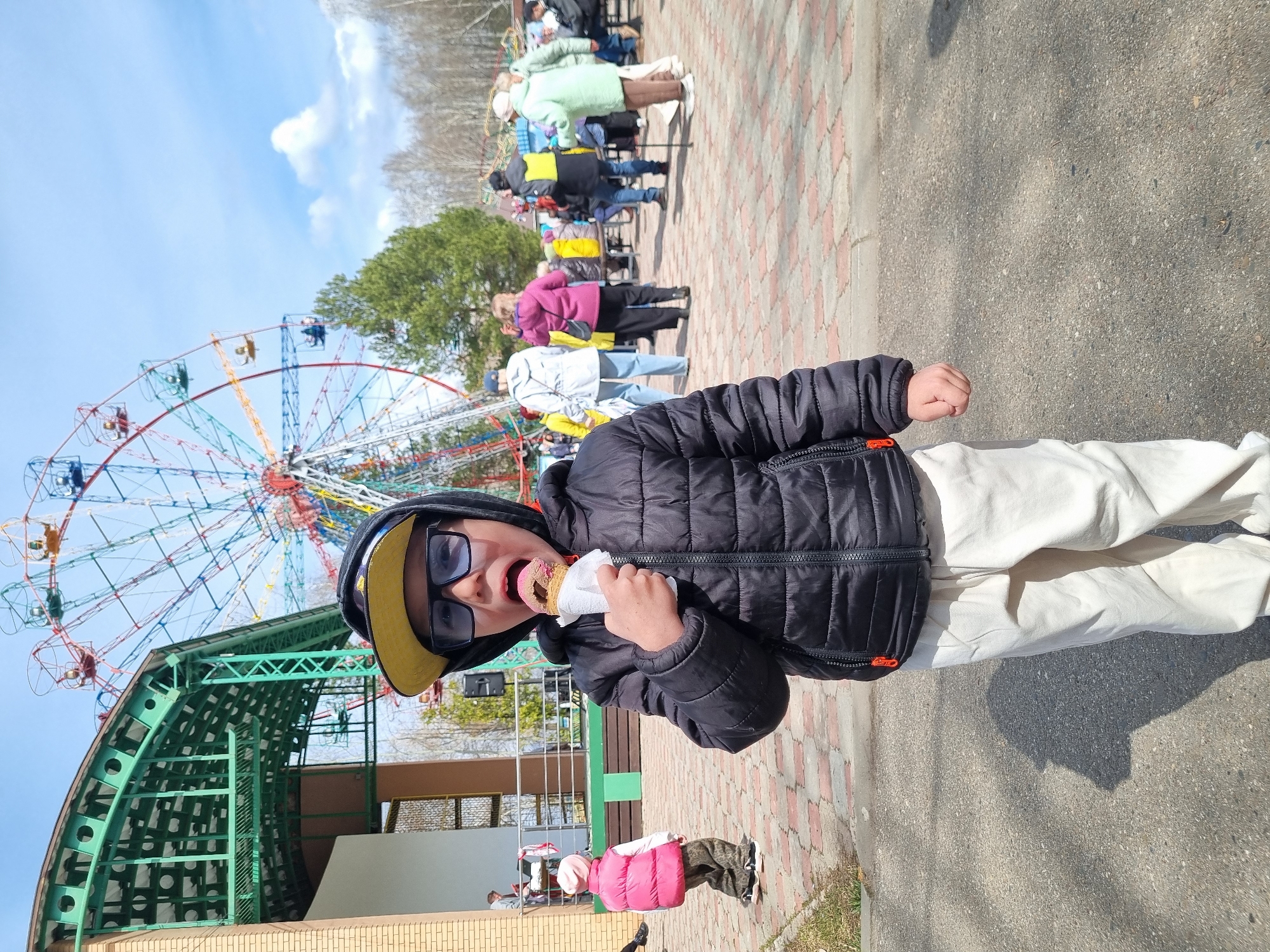This vertical photograph, taken in Chaikovsky, Russia, captures a lively outdoor scene centered around a young boy enjoying an ice cream. The boy, estimated to be between 6 and 8 years old, stands prominently in the foreground, looking directly at the camera with an open-mouthed expression of delight as he holds and eats a pink ice cream cone wrapped in white paper. He is dressed in a black puffer jacket, dark sunglasses, and a yellow baseball cap worn backward, paired with light-colored pants.
The background reveals a bustling amusement park or fairground. A large, colorful Ferris wheel, featuring red, blue, and yellow gondolas, dominates the left side of the frame, towering against a bright blue sky with scattered white clouds. Below the Ferris wheel, a green metal support structure is visible. Several adults are seen in the mid-ground, appearing to queue in a line, suggesting they are waiting for an attraction. Another small child in a pink puffer vest walks away from the camera on the left.
The ground consists of patterned brick pavers and asphalt, with strong shadows indicating a sunny day. Trees with both bare branches and green foliage are visible in the distance, along with parts of buildings. The overall impression is one of enjoyment and recreational activity in a vibrant outdoor setting. No discernible text is visible on signs or clothing.
