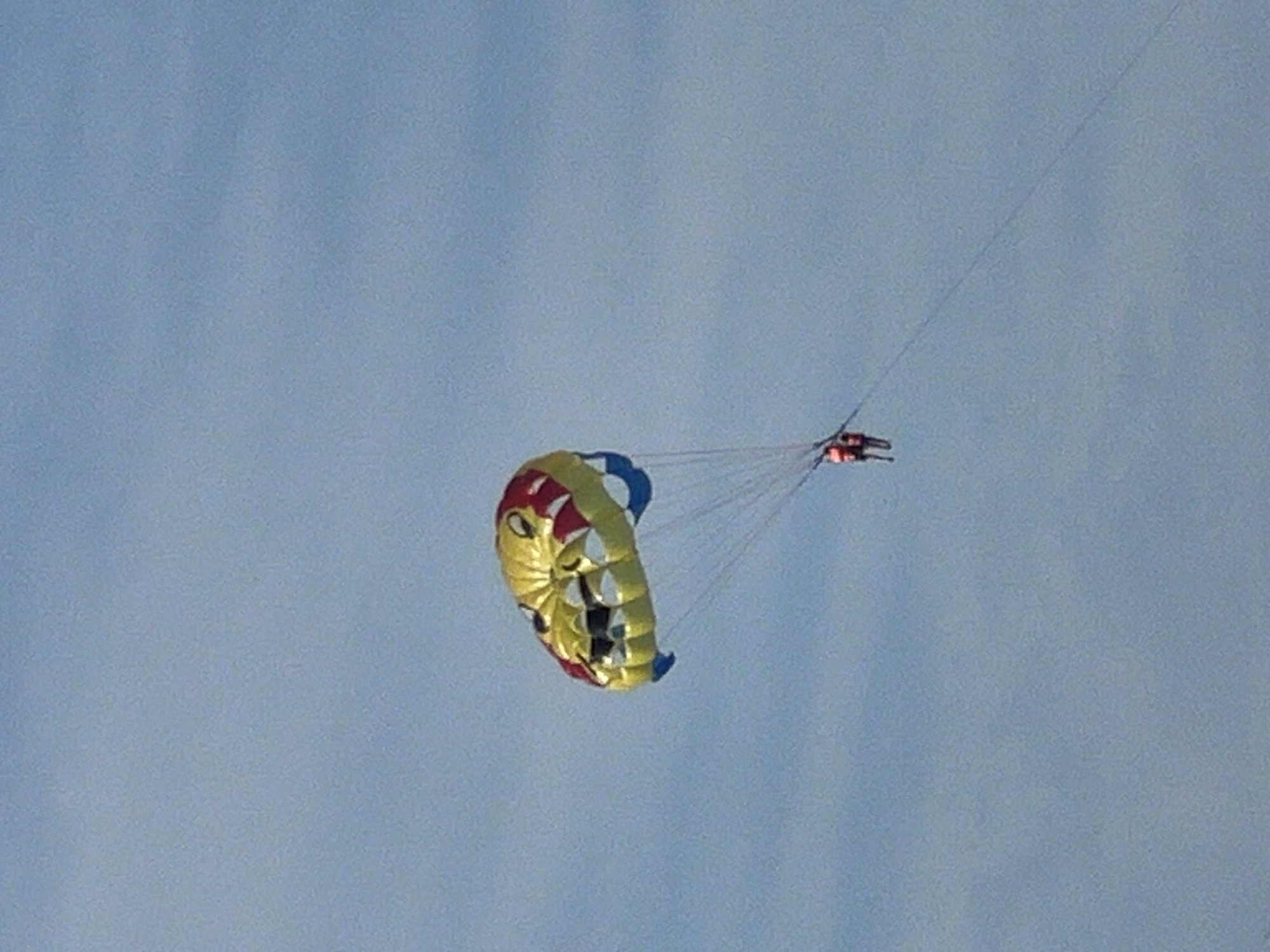 A colorful parasail, designed to resemble a cartoon monster's head with yellow, red, and blue segments, floats against a pale blue sky. Below the parasail, suspended by lines, are two individuals wearing bright orange life vests. They are actively parasailing, their bodies appearing small in the distance against the vast expanse of the sky. The scene is likely from a beach or coastal area in Đà Nẵng, Vietnam, known for its numerous water sports and recreational activities. The overall impression is one of leisure and adventure on a clear day.