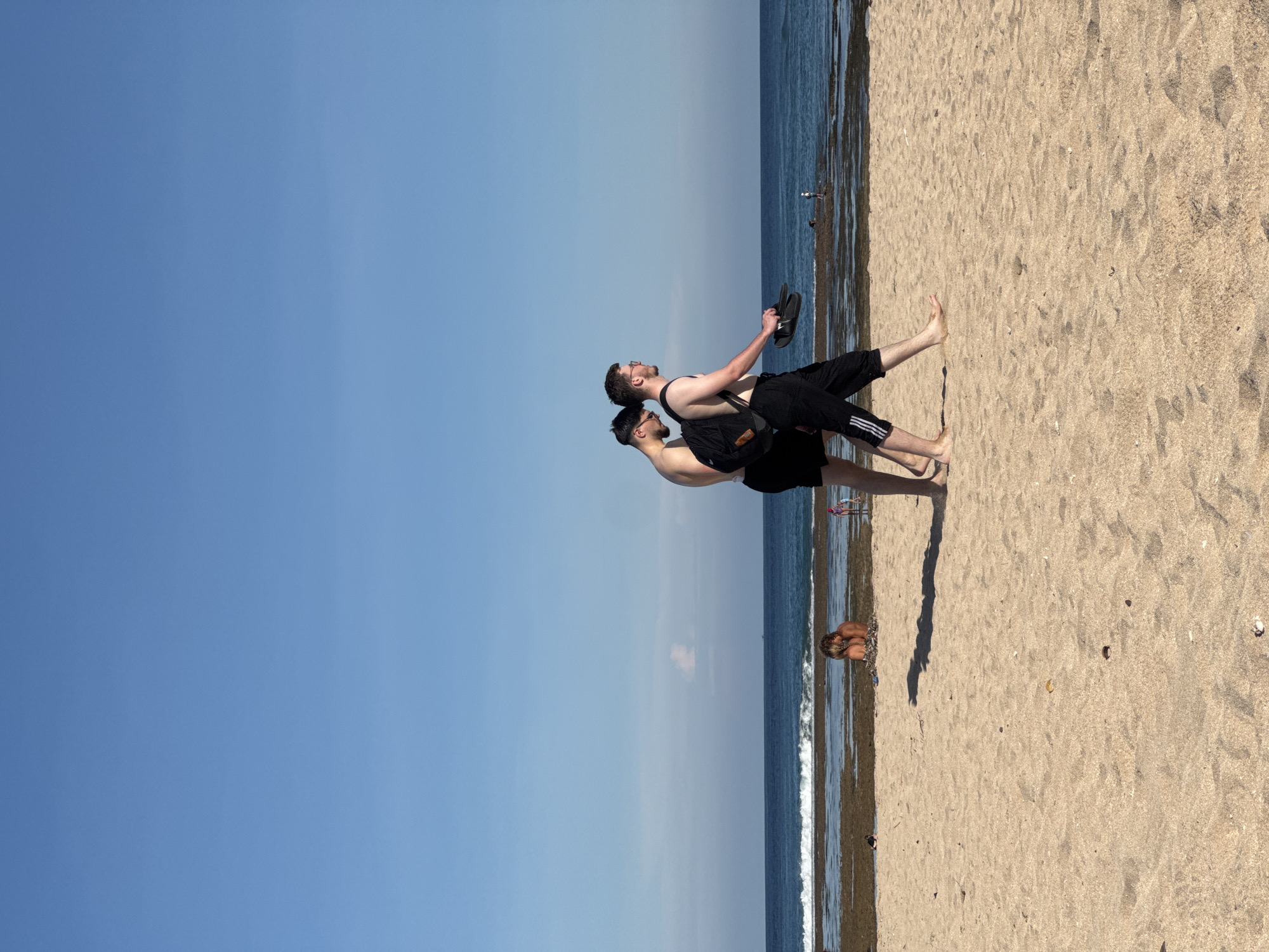 Two men stand on a sandy beach with the ocean on the right and the sky on the left. The man in front wears a black tank top and sunglasses. The man behind him wears a black tank top and glasses, carrying sandals in his right hand and a black bag over his left shoulder. Both men wear black pants. They appear to be interacting in a close embrace. In the background, another individual is crouched near the water's edge. The location is Kuta Selatan, Indonesia.