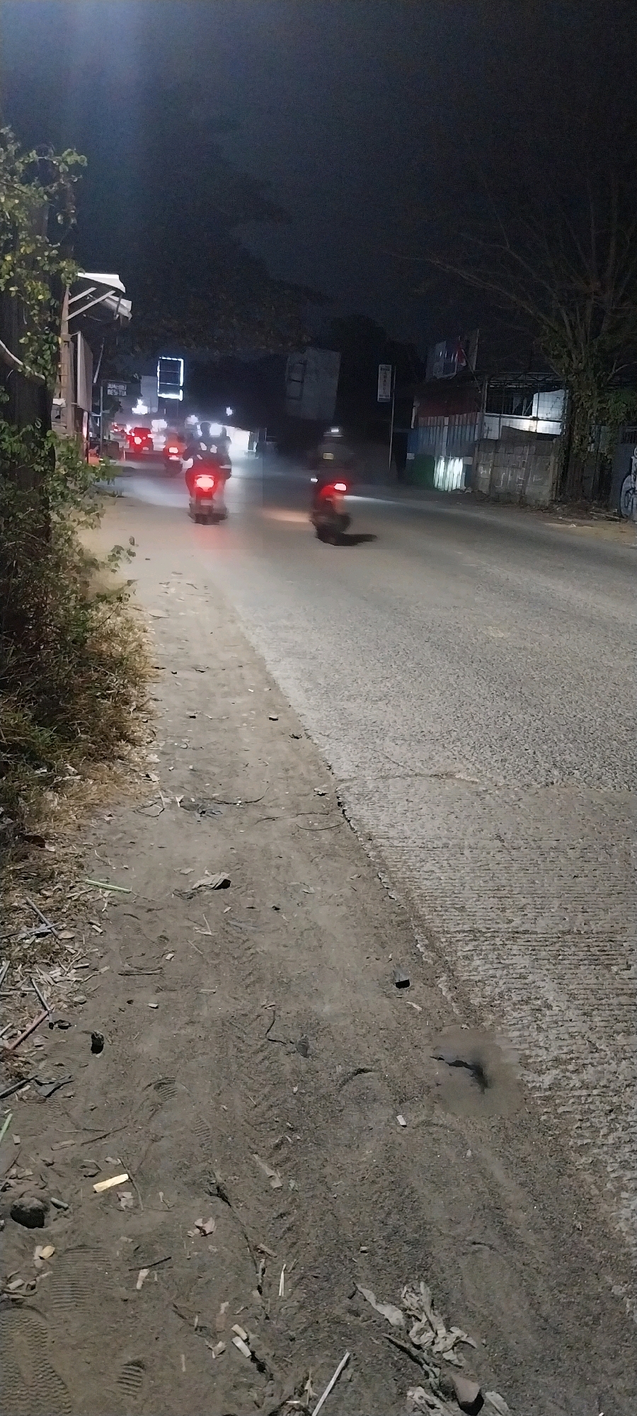 This is a nighttime, long-exposure photograph taken from ground level on a rural road in Kelapa Dua, Indonesia. The scene depicts a road with a dirt shoulder on the left and a paved road on the right. The lighting is low, with the primary illumination coming from the headlights and taillights of vehicles.
Several motorcycles are visible on the paved road, their taillights creating streaks of red light due to the long exposure. One motorcycle is in the foreground, slightly to the right of center, with its red taillight prominently displayed. Further down the road, other motorcycles and at least one car are visible, their lights also blurred by the exposure time.
The dirt shoulder is covered in loose soil, debris, and vegetation. Tire tracks are visible in the dirt, indicating recent vehicle or foot traffic. Scattered pieces of wood, cigarette butts, and other small items are present on the ground.
On the right side of the paved road, buildings and structures are dimly lit. A tree with bare branches stands to the right, silhouetted against the dark sky. The overall impression is one of a quiet, possibly somewhat neglected, rural road at night.
