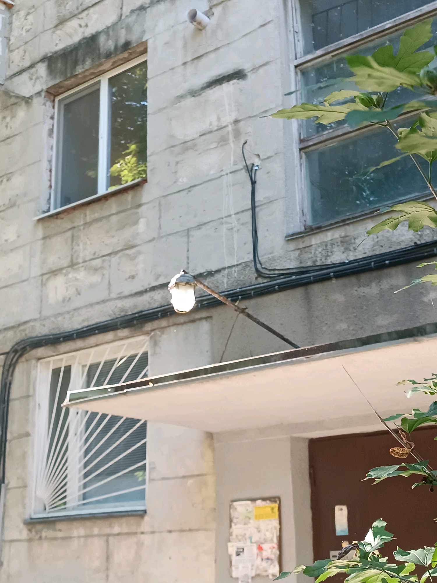 The image depicts the exterior of a Soviet-era apartment building in Chișinău, Moldova. The building's facade is constructed of large, rough concrete blocks, showing signs of wear and some water staining. There are two visible windows on the upper level, one a standard double-pane window and the other a larger window with multiple panes, partially obscured by green leaves from an overhanging tree.
Below the upper windows, a small, illuminated exterior light fixture is mounted on a metal arm extending from the building. Black electrical conduits run along the facade, connecting to the light fixture and other unseen points. Beneath the light, a metal awning extends outward. Below the awning, another window is visible, this one protected by a white metal grate with a radial pattern.
To the right of the awning and grate, a brown door is visible, with a notice board to its left featuring several pinned papers, some of which are yellow. The overall impression is one of a utilitarian, aging urban residential environment. The lighting suggests it is daytime, with bright sunlight casting shadows.