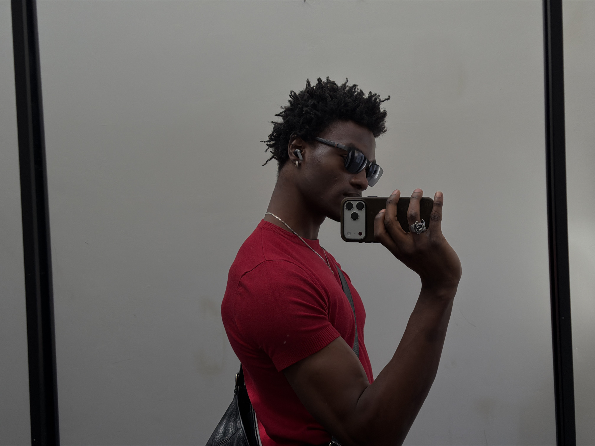 A young Black man is taking a selfie in front of a mirror. He is wearing a red t-shirt, sunglasses, and a silver necklace. He has short, curly hair and is wearing an earpiece in his left ear. He is holding a smartphone in his right hand, which has a large ring on the ring finger. A black bag is slung over his shoulder. The background is a plain gray wall with a black frame on either side of the mirror. The location is not discernible from the image.