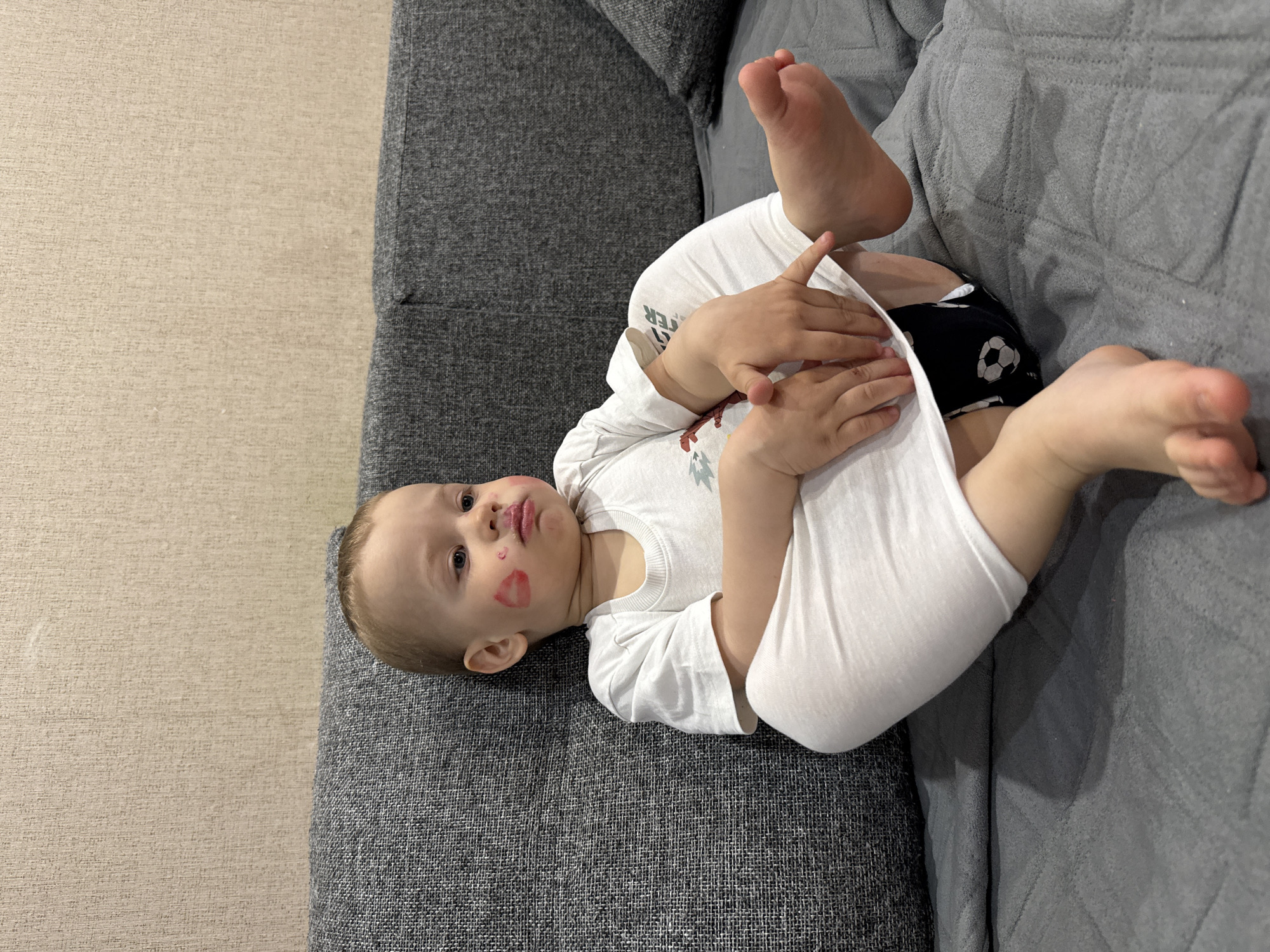 A baby lies on a gray textured couch, possibly in a residential setting, based on the furniture and flooring. The baby has a fair complexion, blonde hair, and blue eyes. Red lipstick marks are visible on the baby's cheeks and mouth. The baby is wearing a white long-sleeved onesie. The baby is wearing black and white soccer-themed shorts. The location is unknown, and Rusanovo, Russia is not confirmed.