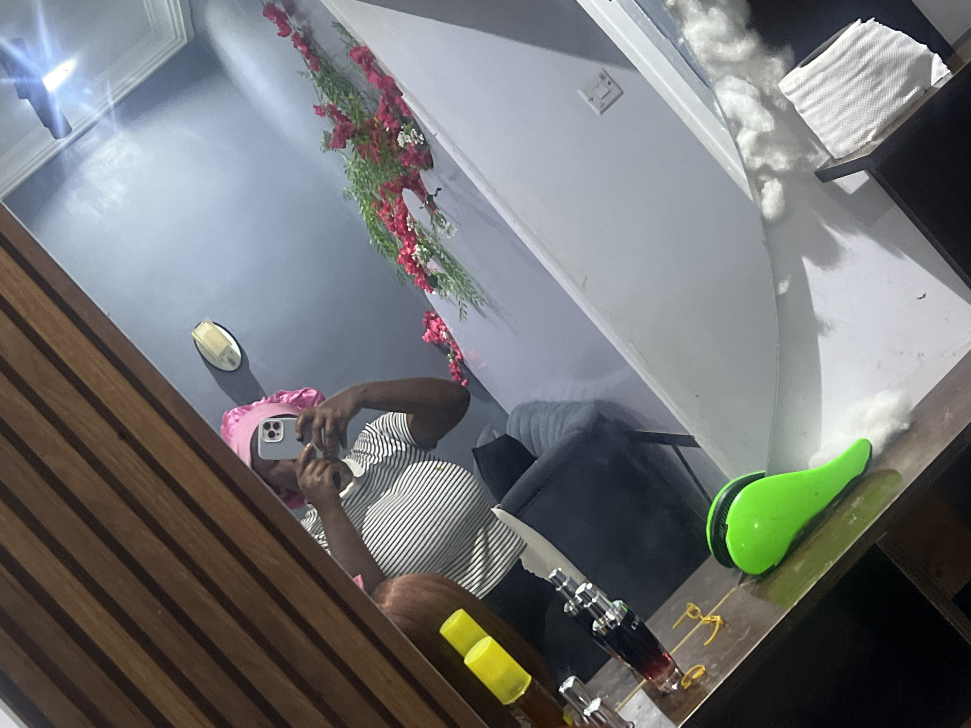 The image depicts a person in Gura Top, Nigeria, taking a mirror selfie. The individual is wearing a pink bonnet and a striped t-shirt, holding a smartphone to capture their reflection. In the foreground, a wooden surface is visible, displaying several items including bottles with yellow caps and a green hairbrush. The background shows a gray wall adorned with a garland of red and white flowers. The lighting suggests an indoor setting, likely a bedroom or dressing area, with a bright light fixture reflecting on the ceiling.
