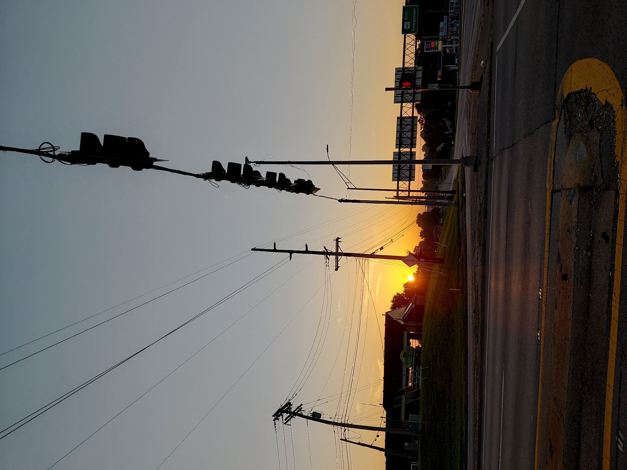 The image captures a sunrise over a commercial area in Frankfort, United States of America. The sky is a gradient of pale blue to soft orange, with the sun just beginning to peek over a grassy hill in the mid-ground. Several power poles with a complex network of wires crisscross the frame, silhouetted against the bright sky. In the foreground, a two-lane asphalt road with yellow dividing lines and a yellow painted curb is visible. On the right side of the road, a green highway sign indicates "460 EAST Georgetown." Beyond the hill, the tops of buildings and trees are visible, with a gas station and convenience store clearly identifiable on the right side. Traffic lights, also silhouetted, hang over the intersection. No people are visible in the scene.