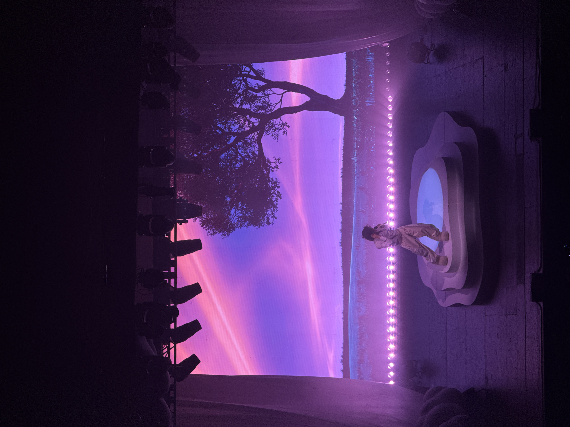 A low-angle, slightly rotated shot depicts a stage scene in what is likely a concert venue. On the right side of the image, a person stands on an elevated, cloud-like platform. To the left, a large screen displays a colorful landscape with a tree and a sunset or sunrise. Stage lighting is visible to the left, indicating a professional production. The location is likely Los Angeles, United States.