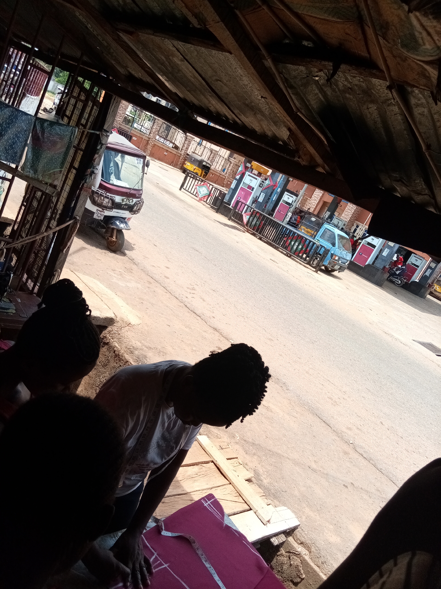 Two dark-haired individuals are seen from the upper torso down, working on a pink cloth with white markings and a measuring tape. They are indoors in what appears to be a shop, with the exterior visible through the shop's opening. The view outside includes a street, a maroon and white three-wheeled vehicle, a blue pickup truck, and a gas station with multiple pink and grey gas pumps. The scene is located in the city of Nsugpe, Nigeria.