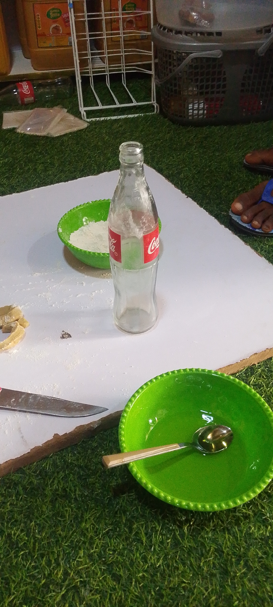 The image depicts a scene in Adankolo, Nigeria. A white surface, possibly a makeshift table, sits on artificial green turf. On the surface, there's an empty Coca-Cola bottle standing upright. A small, green bowl contains white powder. A knife lies next to pieces of dough. Another green bowl contains a spoon and a liquid, possibly oil. In the background, a white wire rack and a grey plastic basket are visible, along with a person's feet wearing blue sandals. A bottle of oil is partially seen in the background.