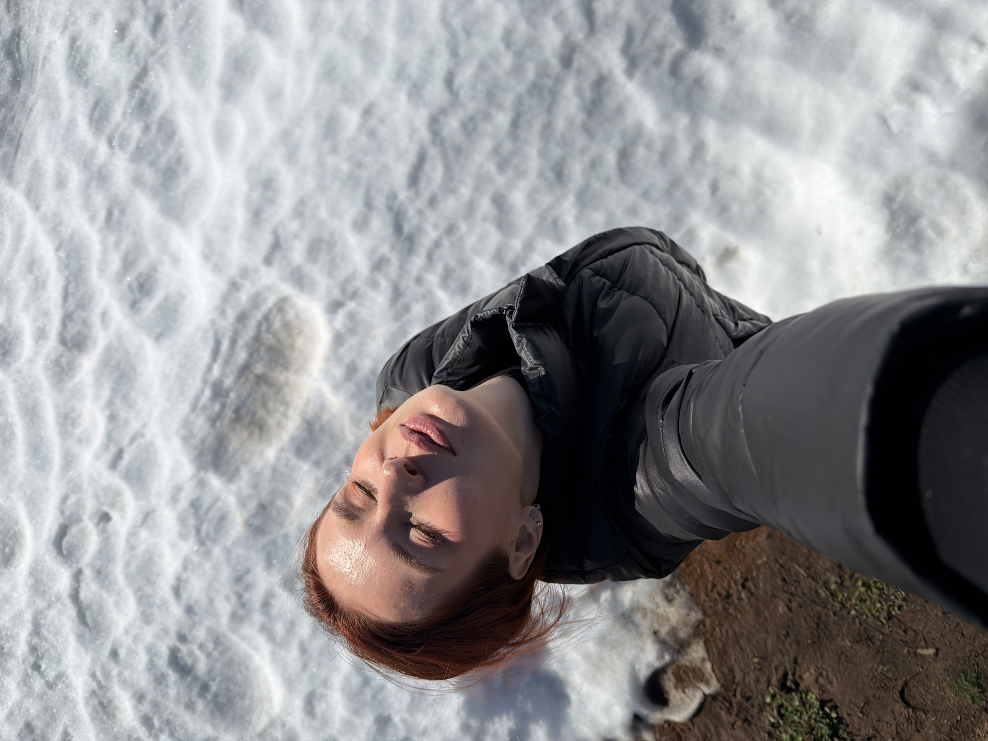 The image presents an overhead selfie of a woman in an outdoor, snowy environment within the city of Chaikovsky, Russia. The woman, possessing reddish-brown hair, wears a black puffer jacket. Her head is tilted upward and slightly to her right, with her face directed towards a strong light source, causing bright highlights on her skin, which exhibits a noticeable sheen on her forehead and cheeks. Her eyes are closed, and her lips are slightly parted. She is wearing multiple small hoop earrings on her visible left ear. The background is dominated by white, textured snow, displaying various ripples and depressions. In the lower-left section of the frame, areas of bare, dark brown earth are visible, interspersed with sparse green vegetation, indicating partial snowmelt. The strong sunlight creates a high contrast scene with bright snow and illuminated facial features. The perspective suggests the image was captured by the woman herself, extending her arm forward and downward.