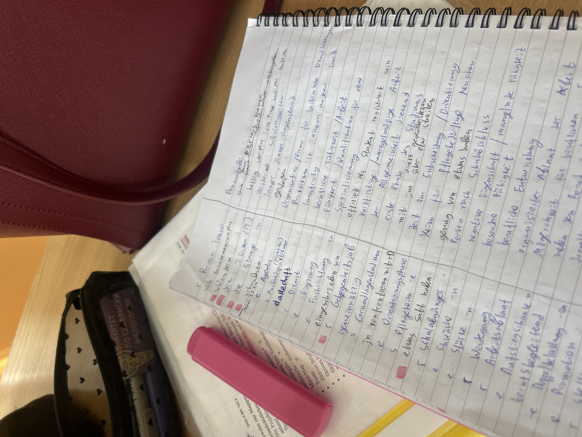 A notebook is open on a table, filled with handwritten notes in German. Beside it, there is a pen case, a pink highlighter, and some printed documents. The notes appear to be study notes.