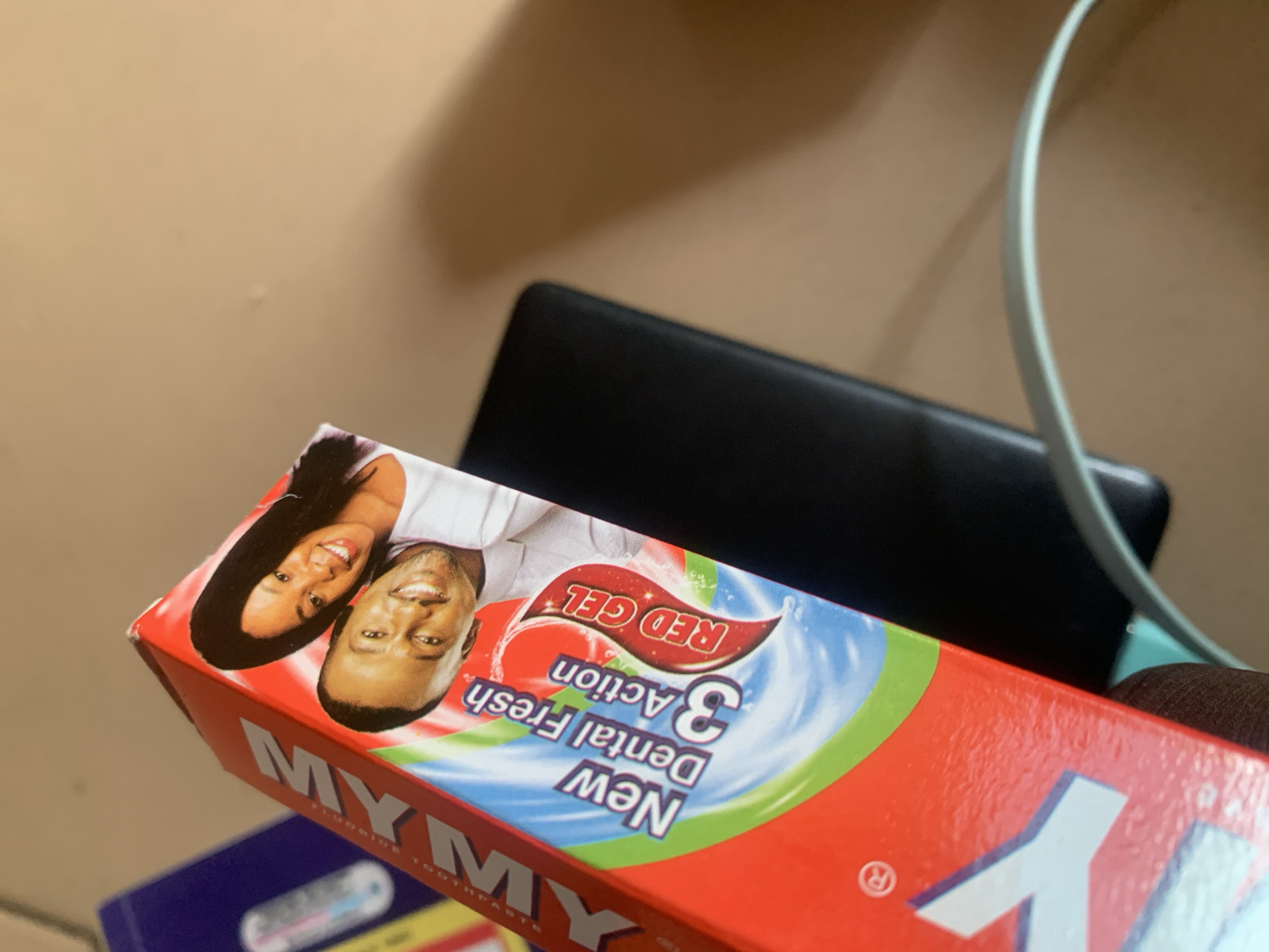 This is a close-up, angled shot of a red toothpaste box. The box has a white logo that says "MY..." and below that, "New Dental Fresh 3 Action RED GEL". There is a picture of a smiling Black couple on the box. The box is leaning against a black object, possibly a phone, and a blurry, light blue cable is visible in the foreground. The background appears to be a tan wall. There are no people visible in the city of Jauro Yinu, Nigeria in this image.