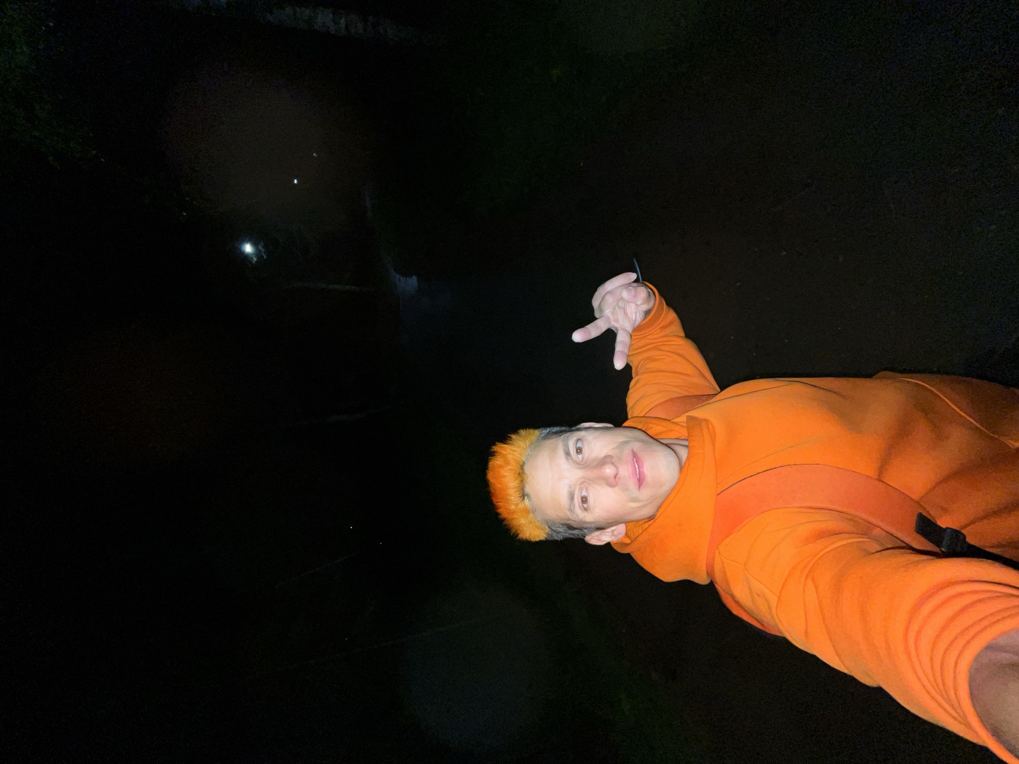 The image depicts a male individual taking a selfie at night in Chaikovsky, Russia. The man is positioned centrally, wearing a bright orange hooded sweatshirt with orange backpack straps visible. He has short, bright orange hair styled upwards and a light complexion. His right hand is raised, performing a 'V' gesture with the index and middle fingers extended; a thin, dark object is visible between his middle and ring fingers. The background is largely dark, revealing a pathway or road bordered by dark tree silhouettes. Two distinct streetlights are visible in the mid-ground on the right side of the path, with a third, fainter light source further in the distance, providing minimal illumination to the scene.