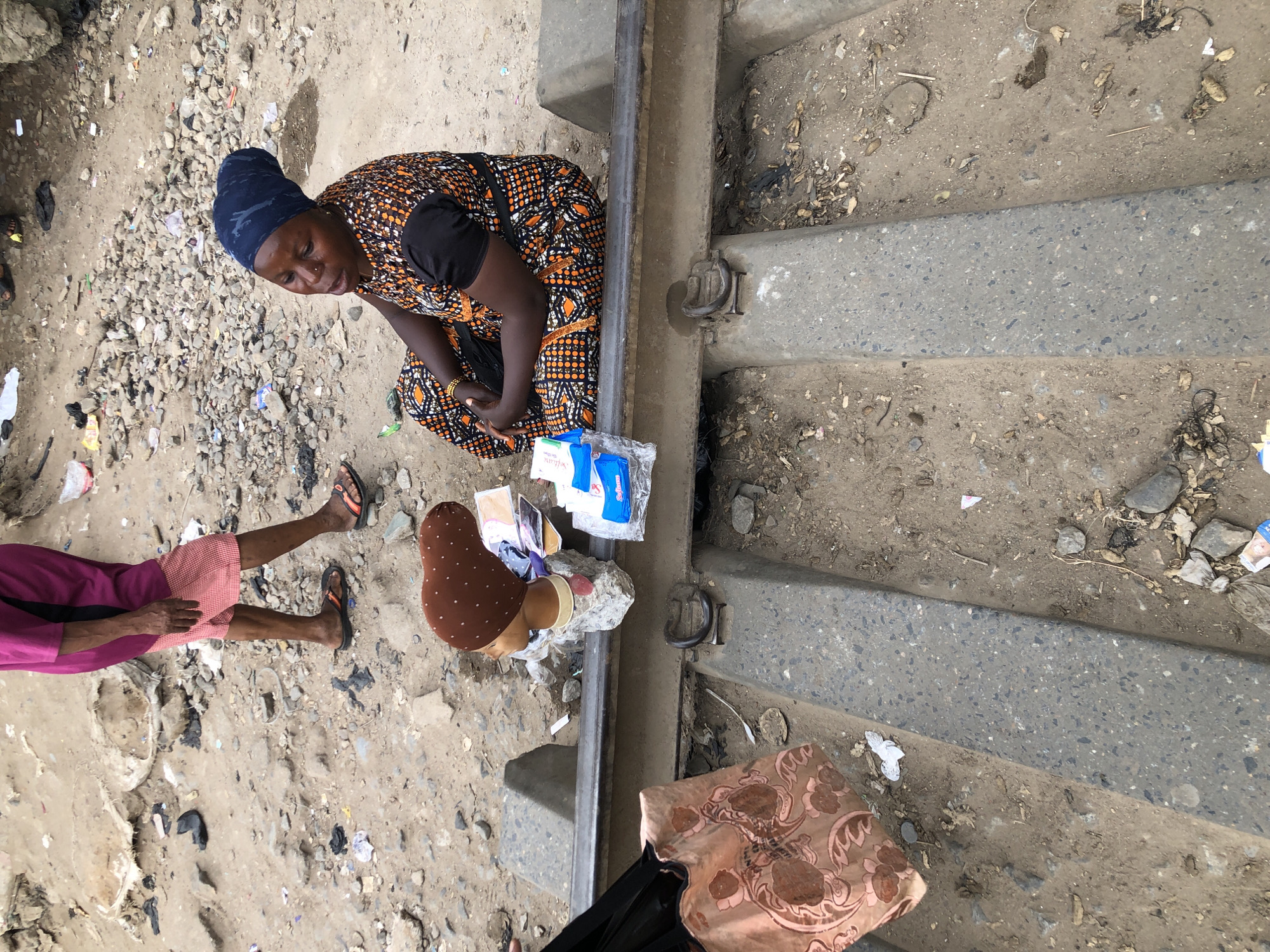 In an outdoor scene in Lagos, Nigeria, a woman sits on a railway track, her hands clasped in her lap, wearing an orange and black patterned dress and a dark blue headscarf. Next to her, on the same track, is a mannequin head displaying a brown headscarf adorned with small white dots. A pink and brown patterned bag lies on the ground to the left of the mannequin. To the right of the woman, a stack of what appears to be sanitary pads is visible. In the background, another person, partially visible from the waist down, stands wearing a magenta tunic and red and white checkered shorts. The ground surrounding the railway is rough and littered with debris, including rocks, dirt, and plastic waste.